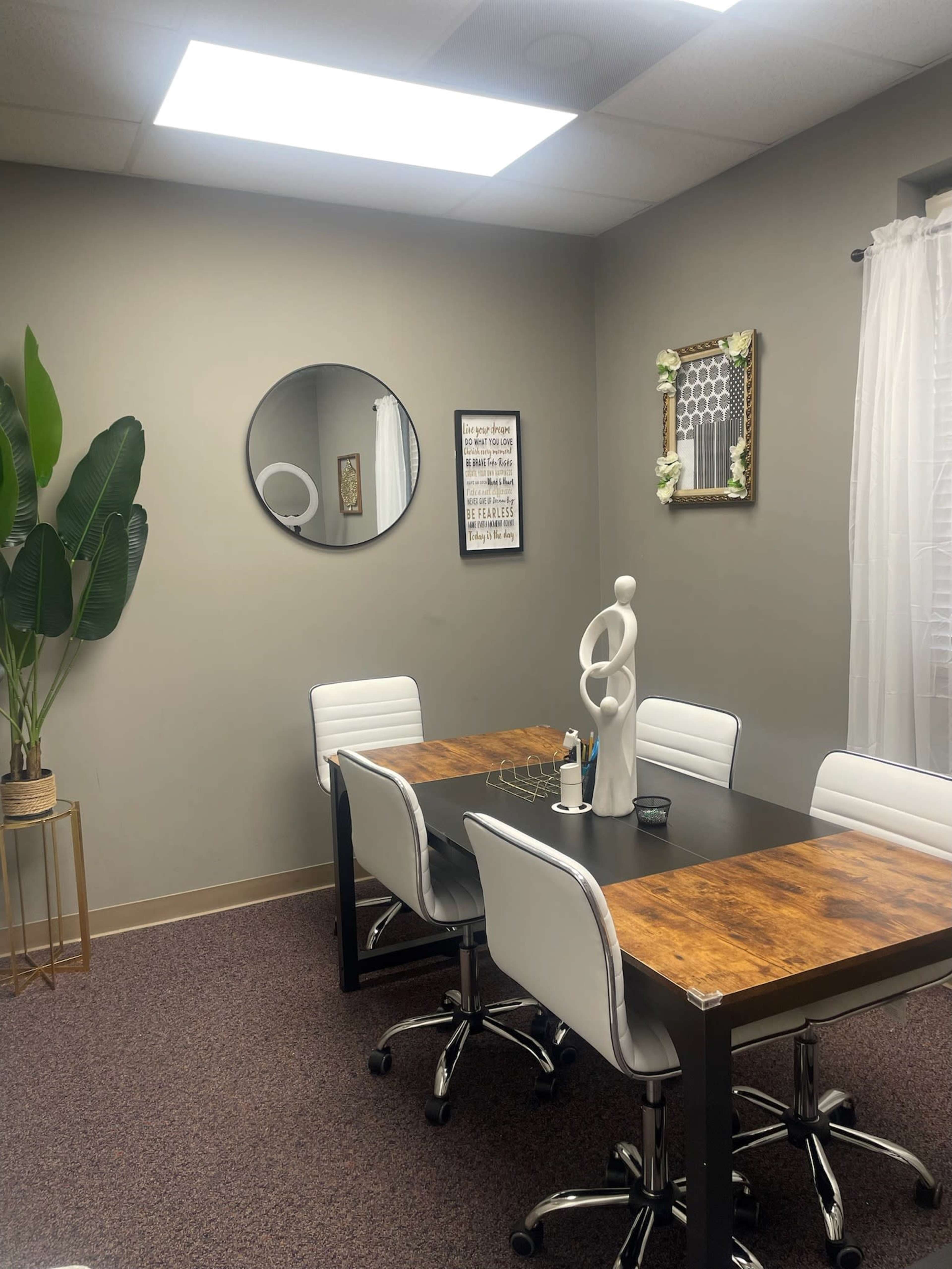 A small conference room features a wooden table surrounded by white chairs, with a large mirror, decorative art, and a potted plant on the wall.