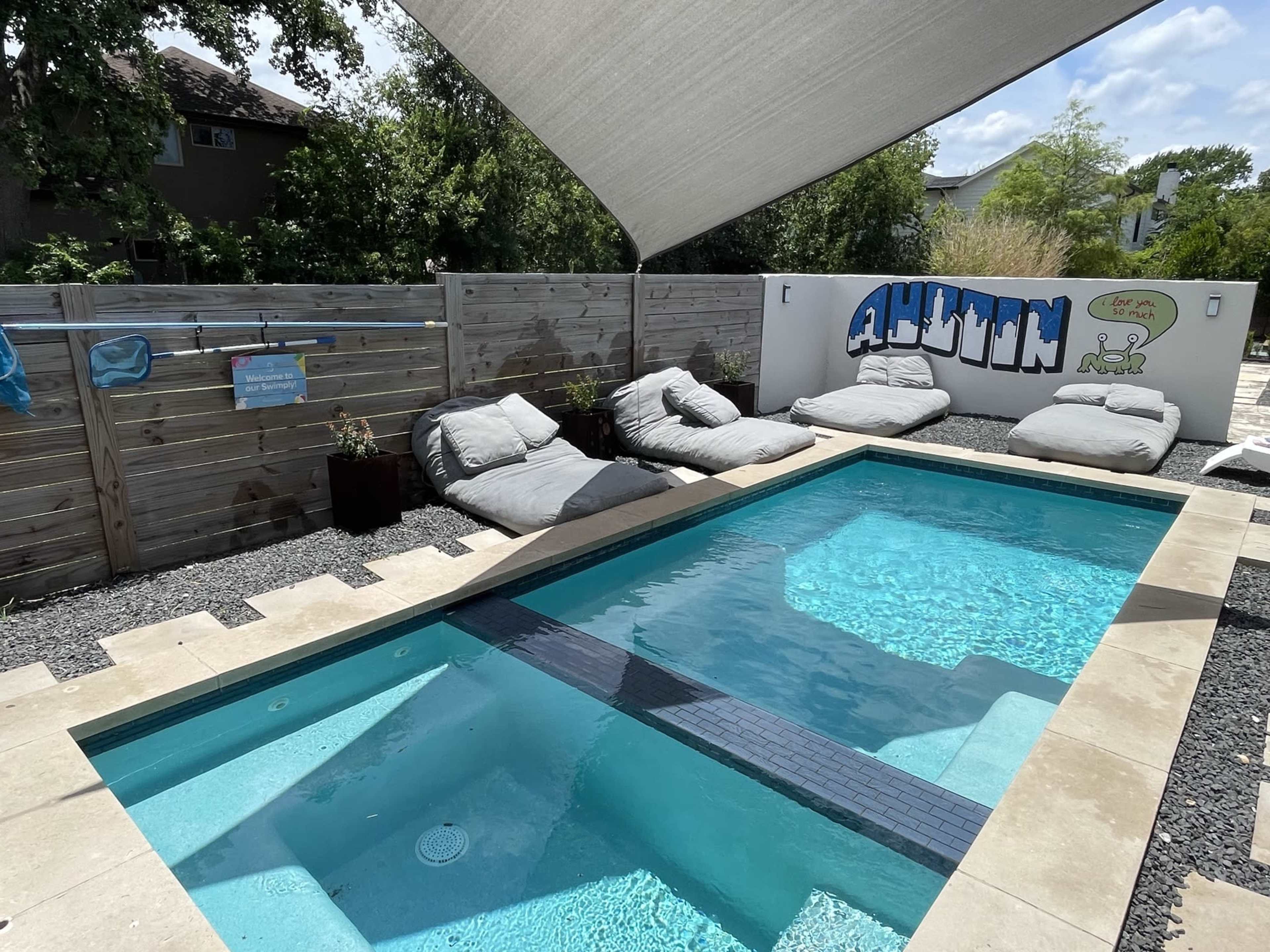 A rectangular swimming pool with a light stone deck is surrounded by large lounge cushions and a graffiti-style mural on a wooden fence.