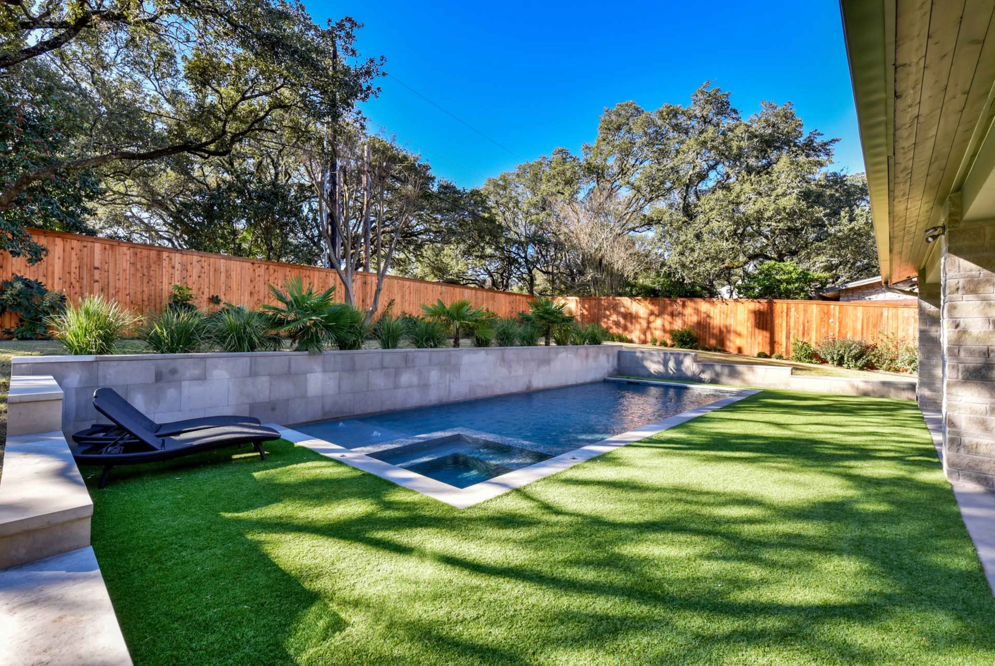 The image shows a modern backyard with a rectangular swimming pool surrounded by artificial grass and a wooden fence.