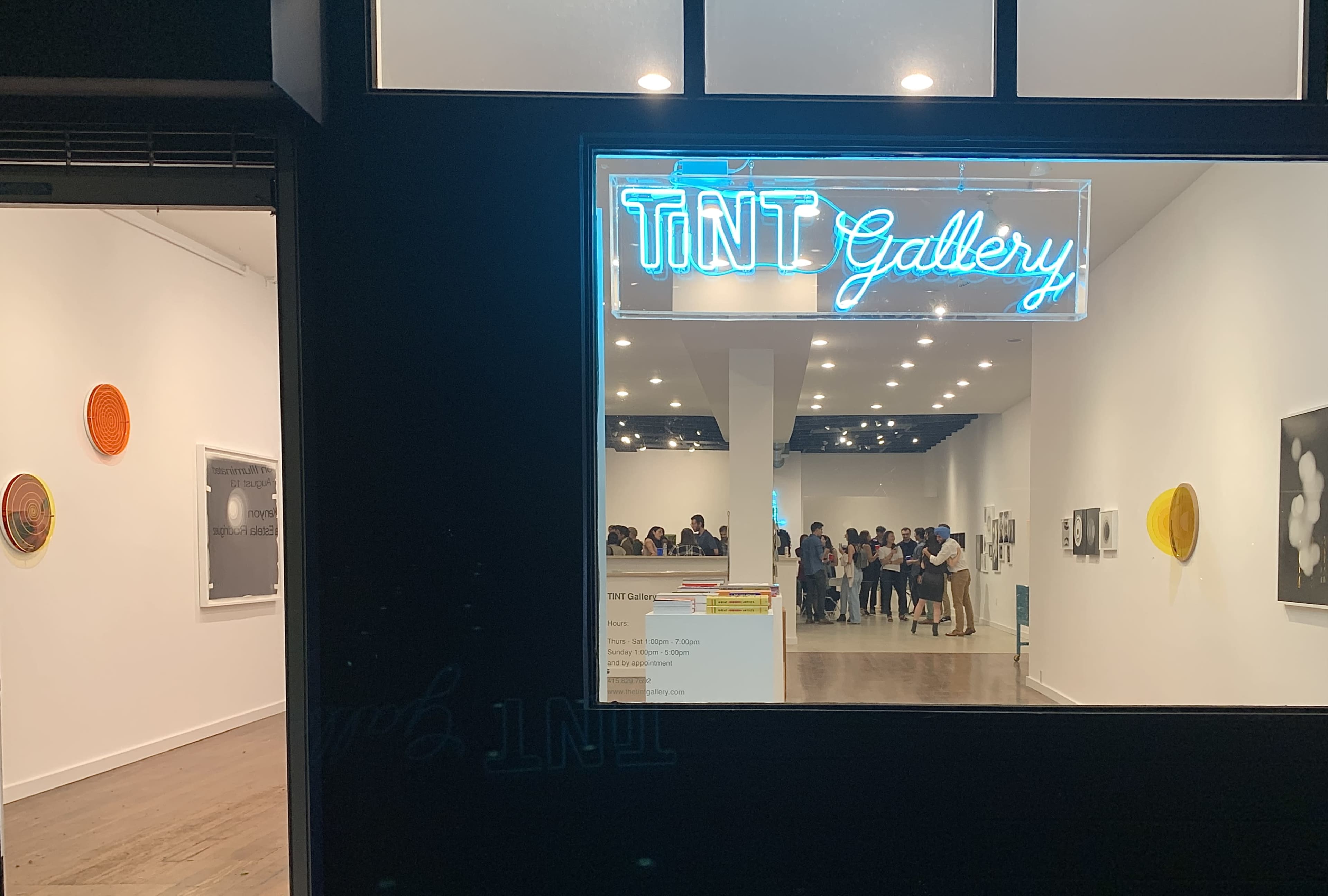 The image shows the entrance of TNT Gallery, featuring a neon sign and visitors gathered inside.