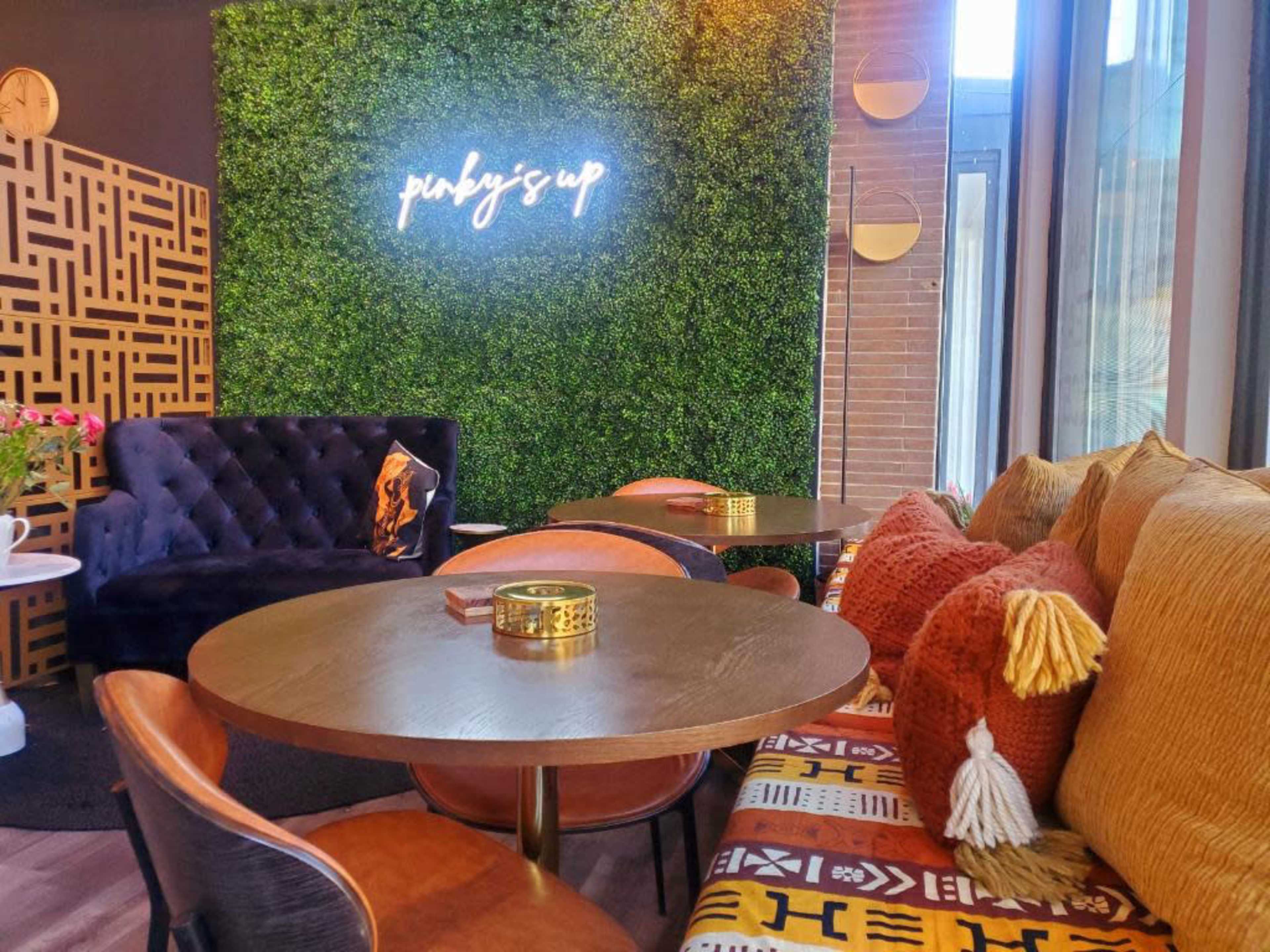 A cozy seating area with a dark blue sofa, orange and patterned cushions, and a green wall featuring the text "pinkys up" in neon lights.