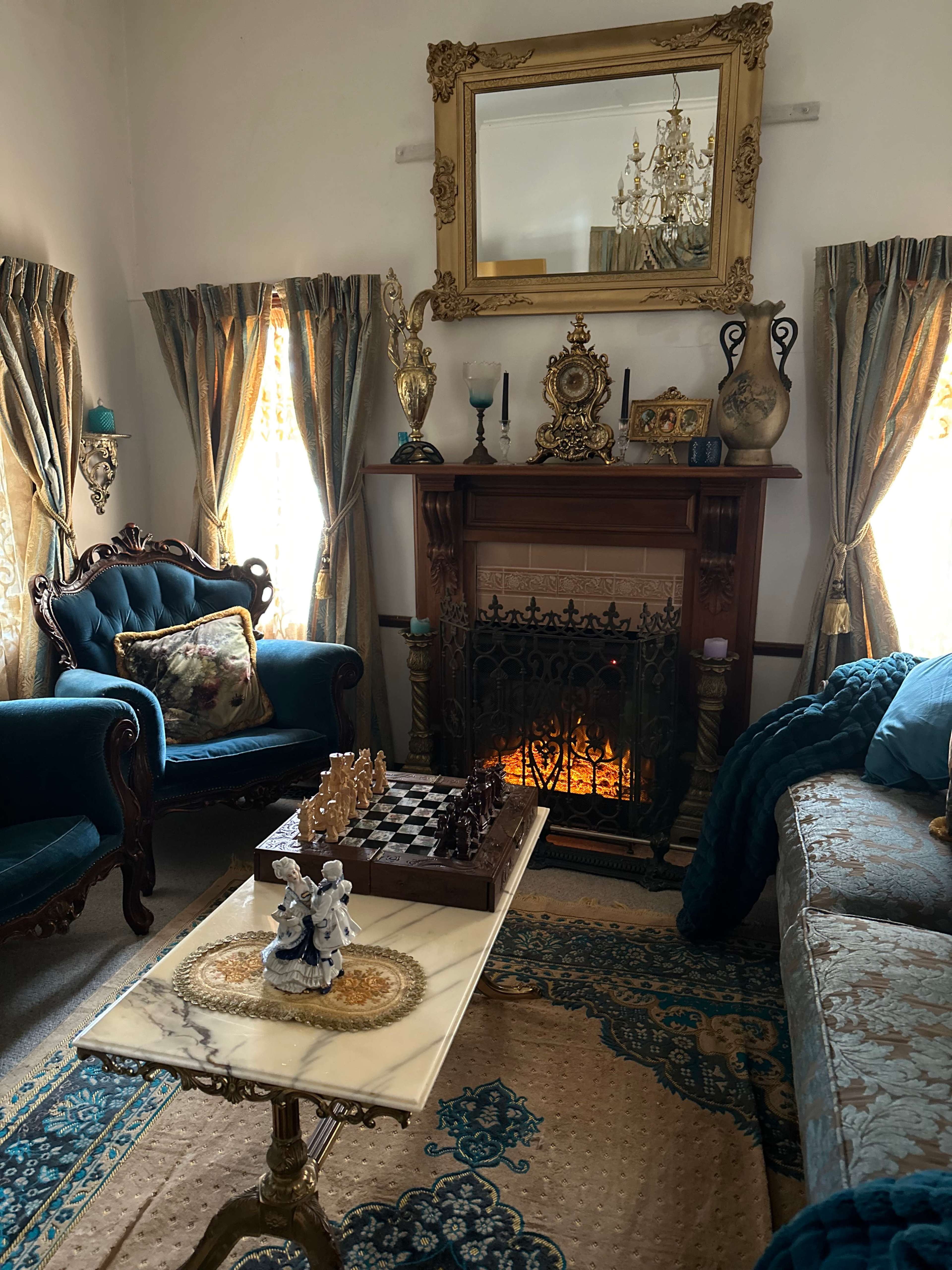 A cozy living room with a fireplace, elegant furniture, a chess board on a table, and ornate curtains framing the windows.