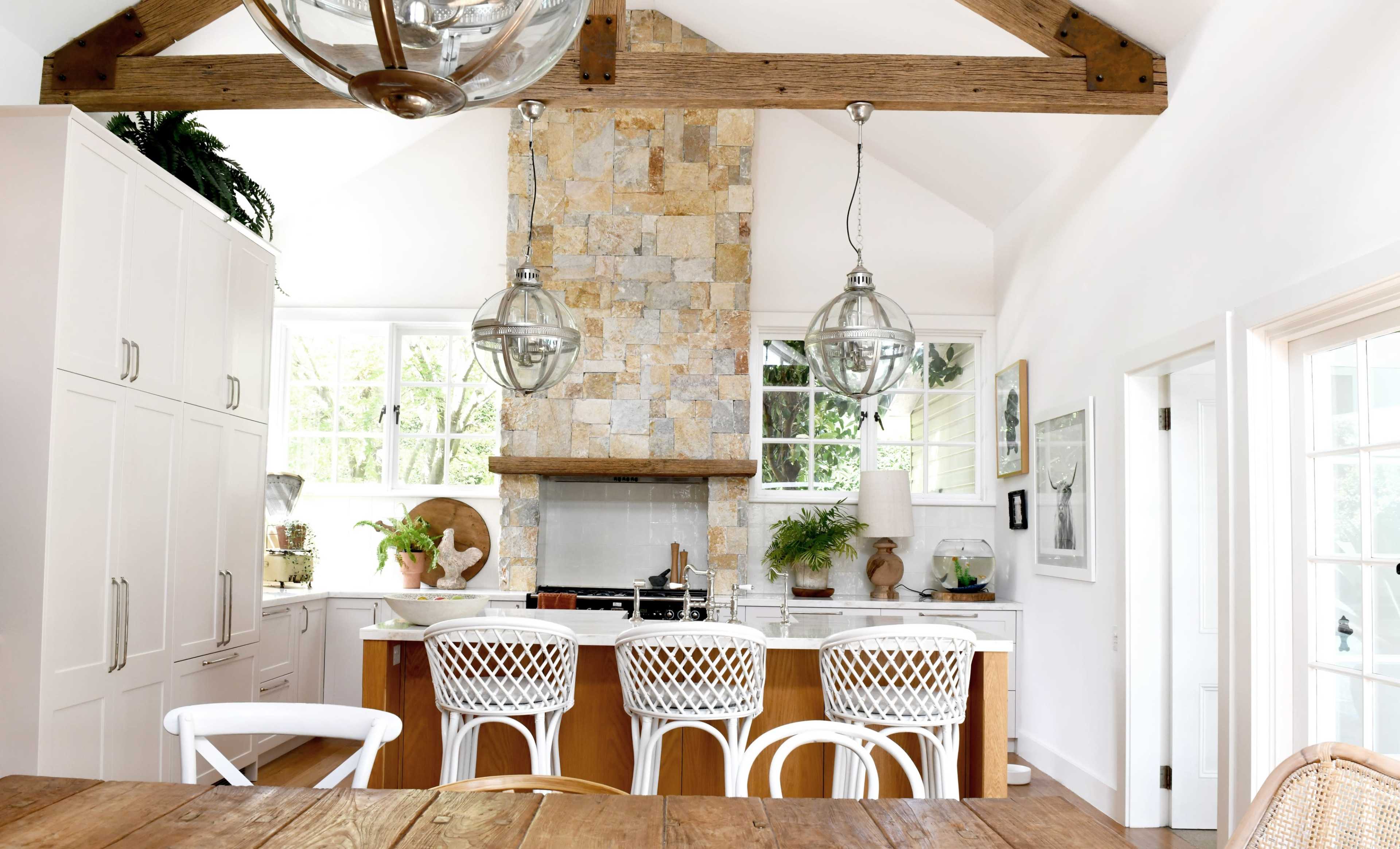 The image depicts a modern kitchen with a stone accent wall, white cabinetry, and four white barstools arranged around a wooden island.