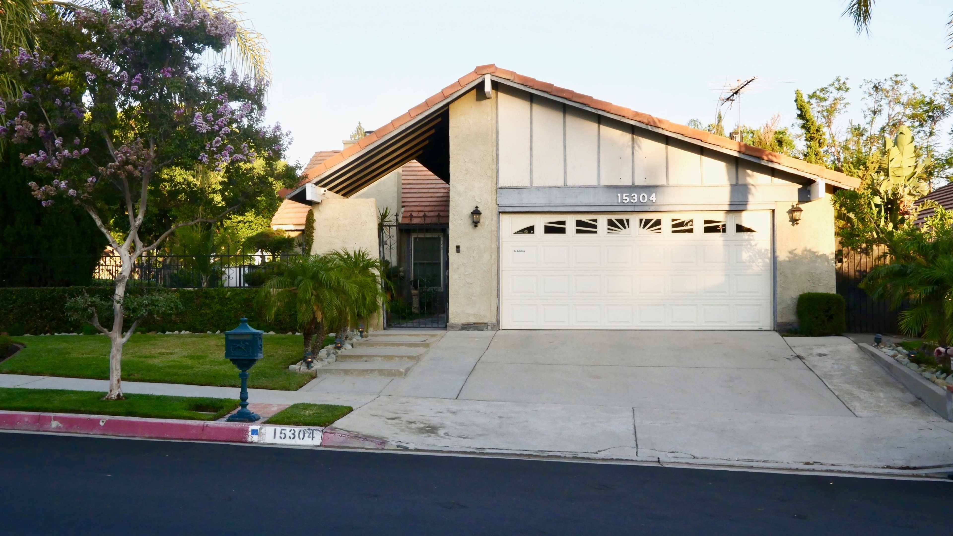 A single-story house with a two-car garage sits on a well-kept lawn surrounded by trees and shrubs.