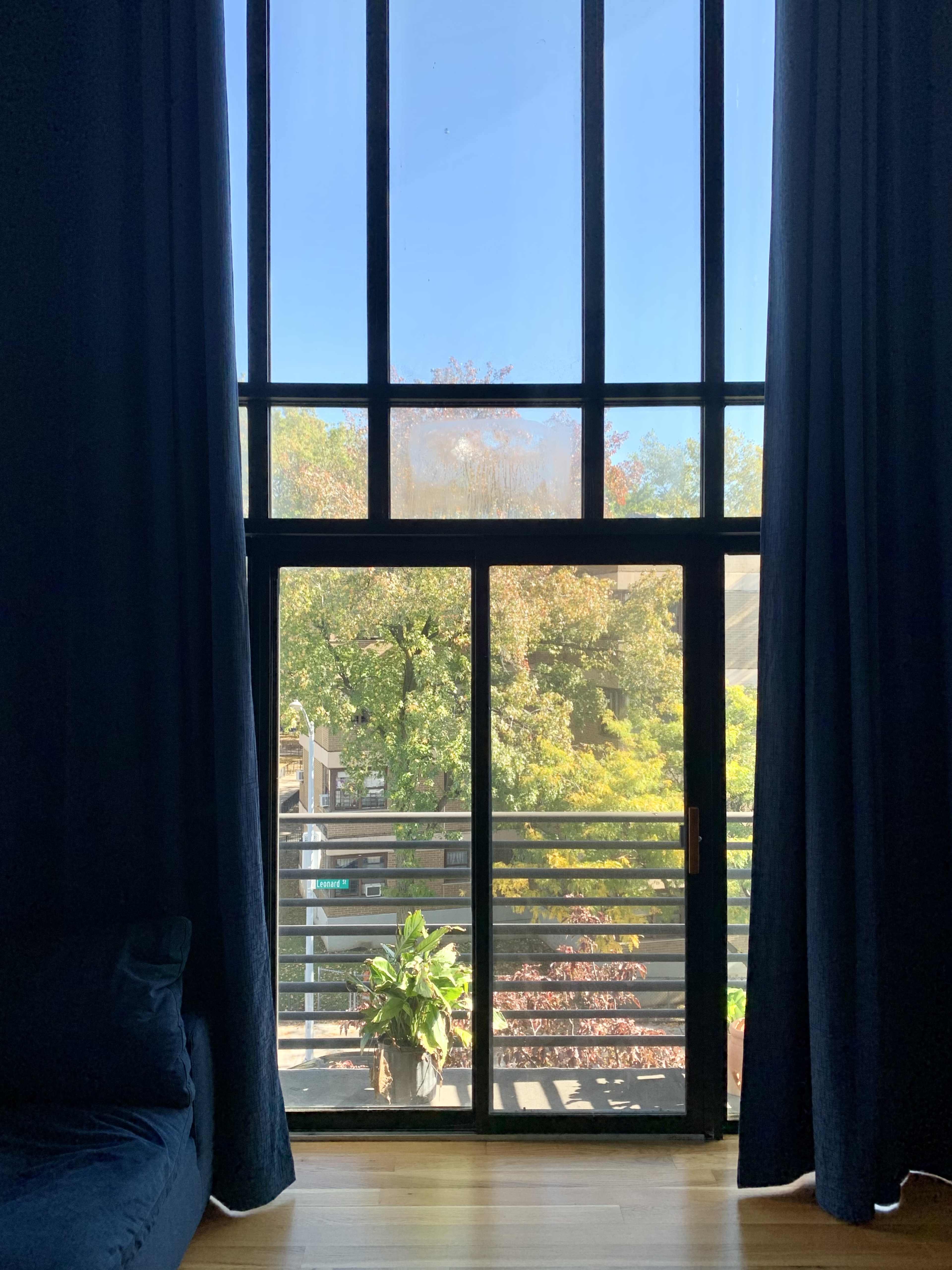 A tall window with black frames and sheer curtains overlooks a balcony with greenery outside.