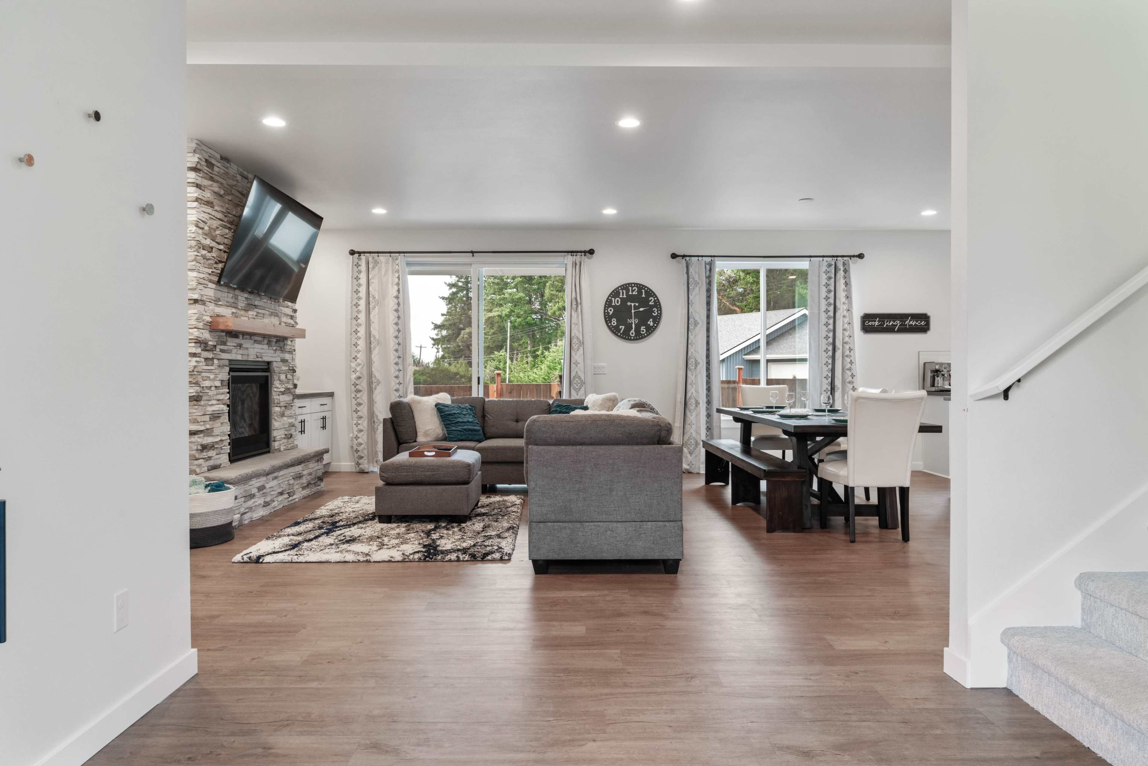 A modern living space with a gray sectional sofa, a television mounted on the wall, and a dining area with a table and chairs.