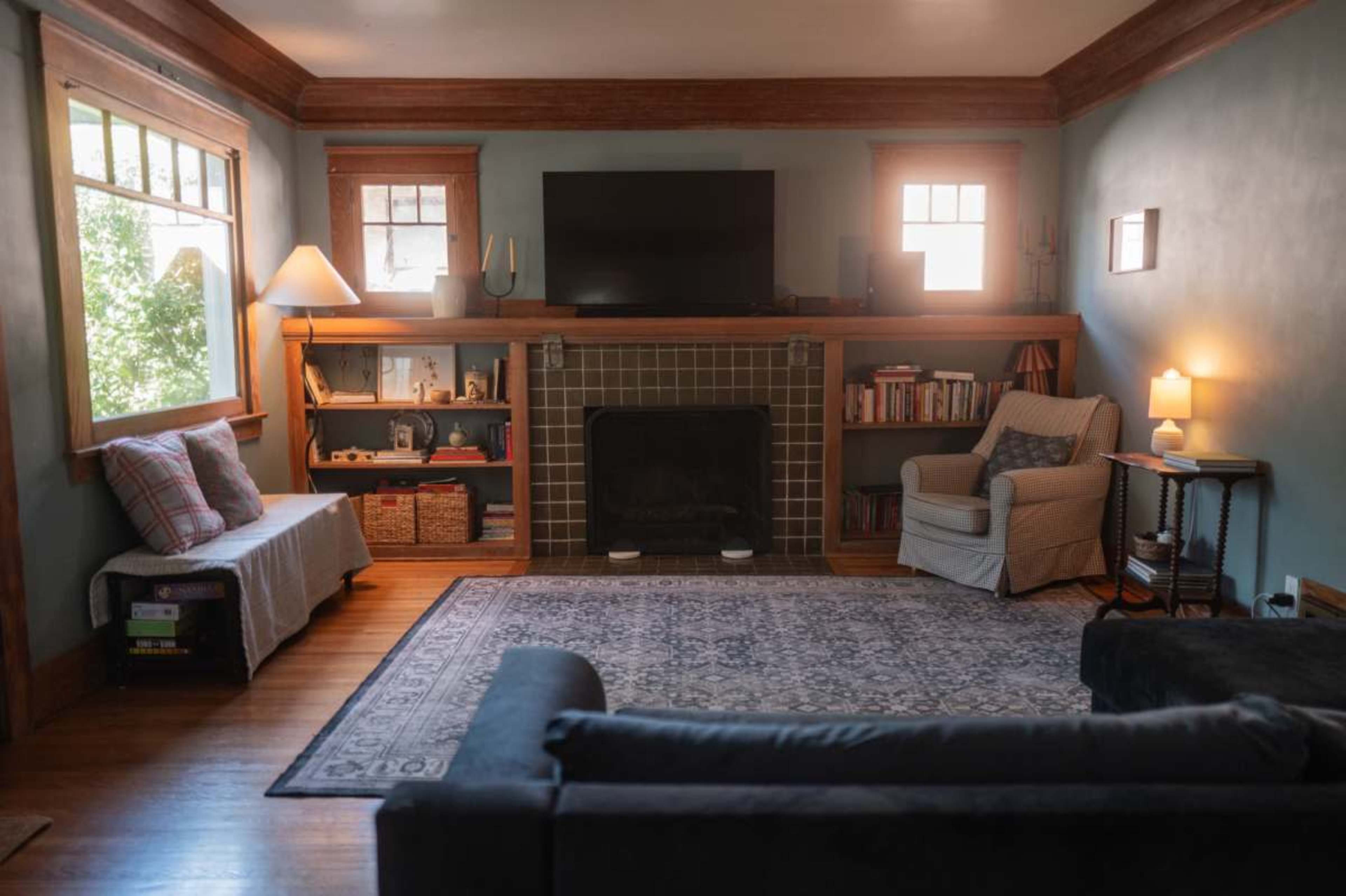 The image shows a cozy living room with a television above a mantel, a patterned area rug, and comfortable seating areas.