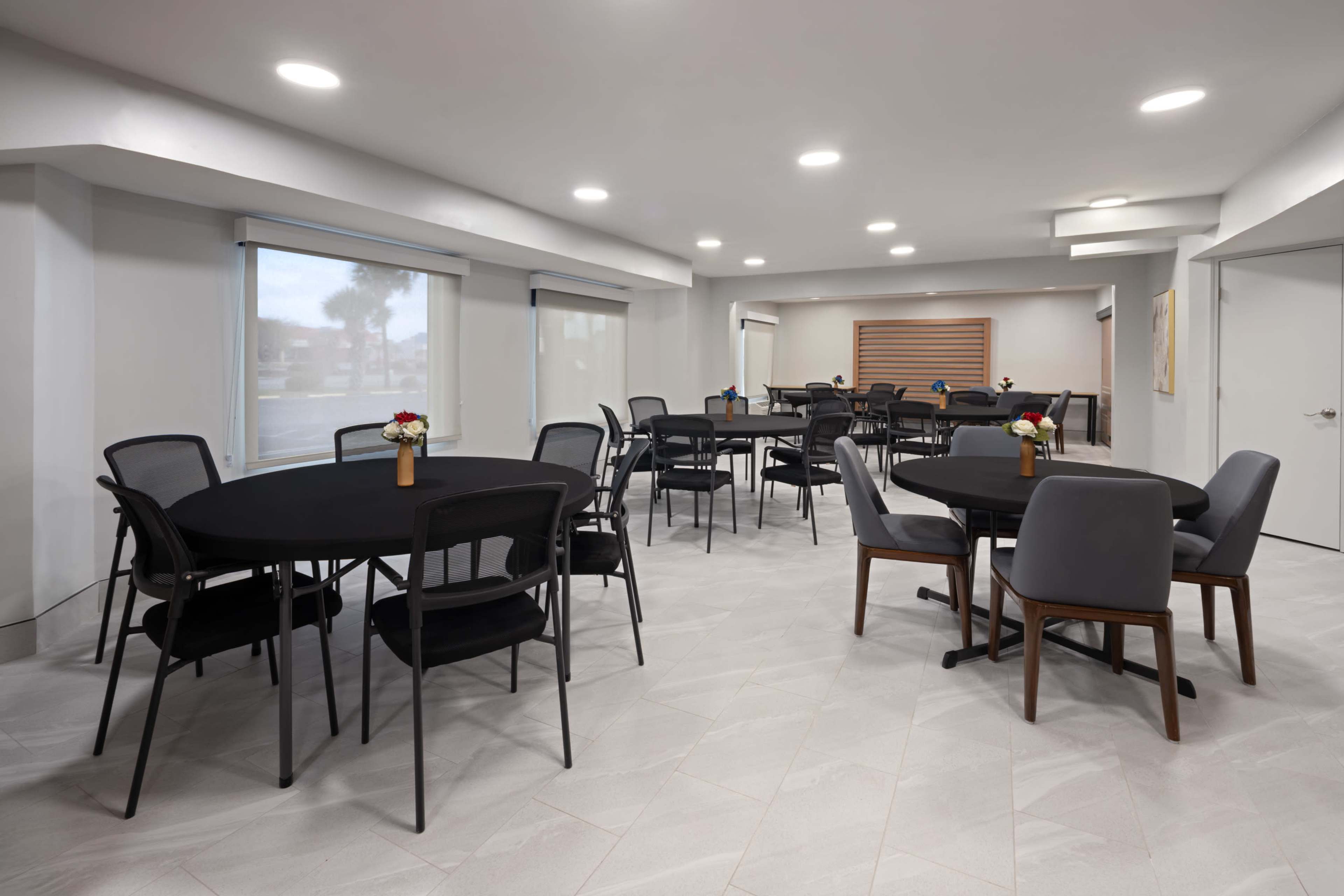 The image shows a modern event space with several round tables and chairs arranged around a light-colored tiled floor, illuminated by overhead lights.