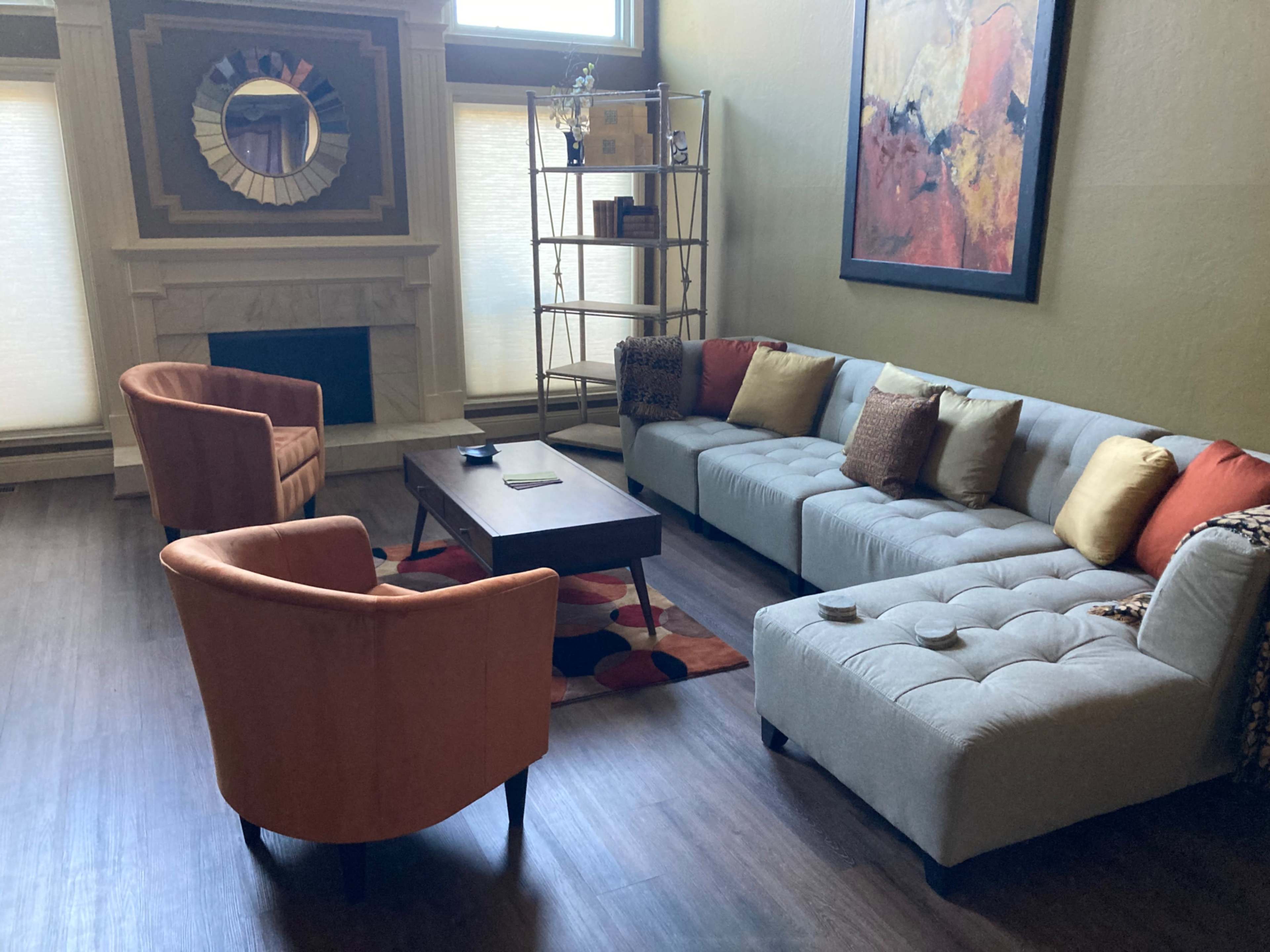 A living room with a gray sectional sofa, two orange armchairs, a wooden coffee table, and a bookshelf against a wall, all arranged around a fireplace.