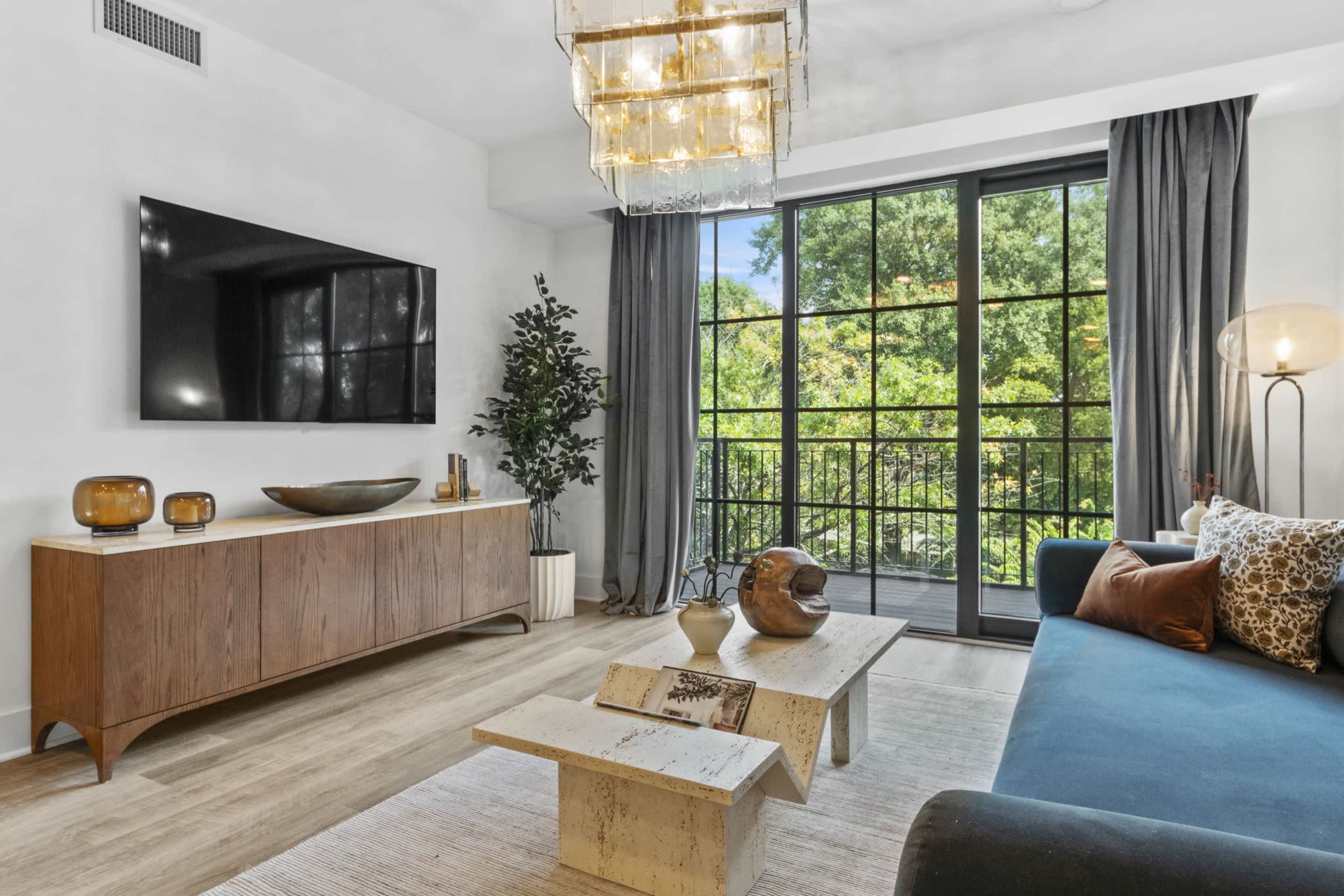 The living room features a modern design with a television mounted on the wall, a wooden sideboard, a large window showcasing greenery outside, and a combination of seating and decorative elements.