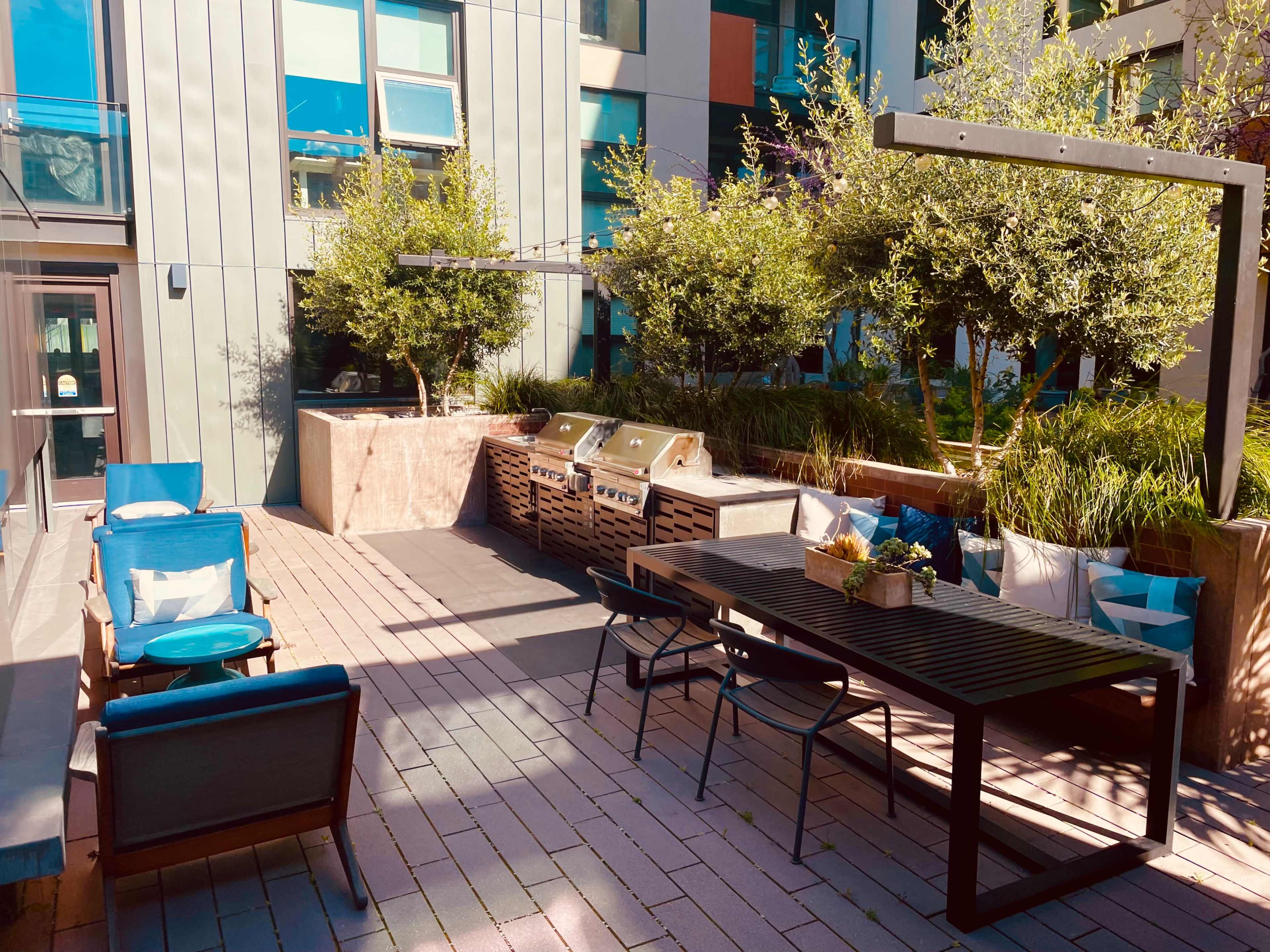 A modern outdoor patio area with BBQ grills, a dining table, and seating surrounded by greenery and built structures.