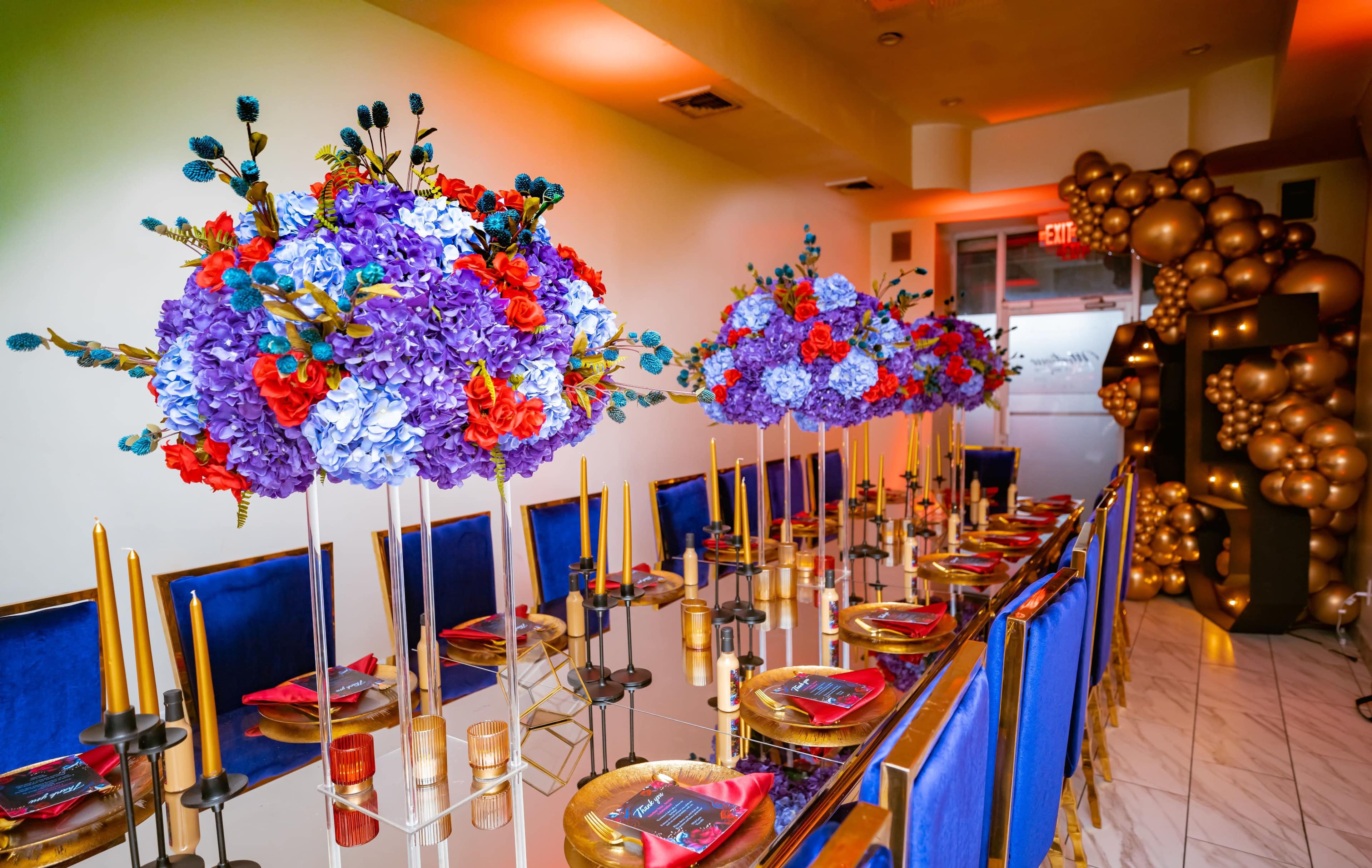 A long dining table is adorned with tall floral arrangements, golden candlesticks, and styled dinnerware, set in a brightly lit room.