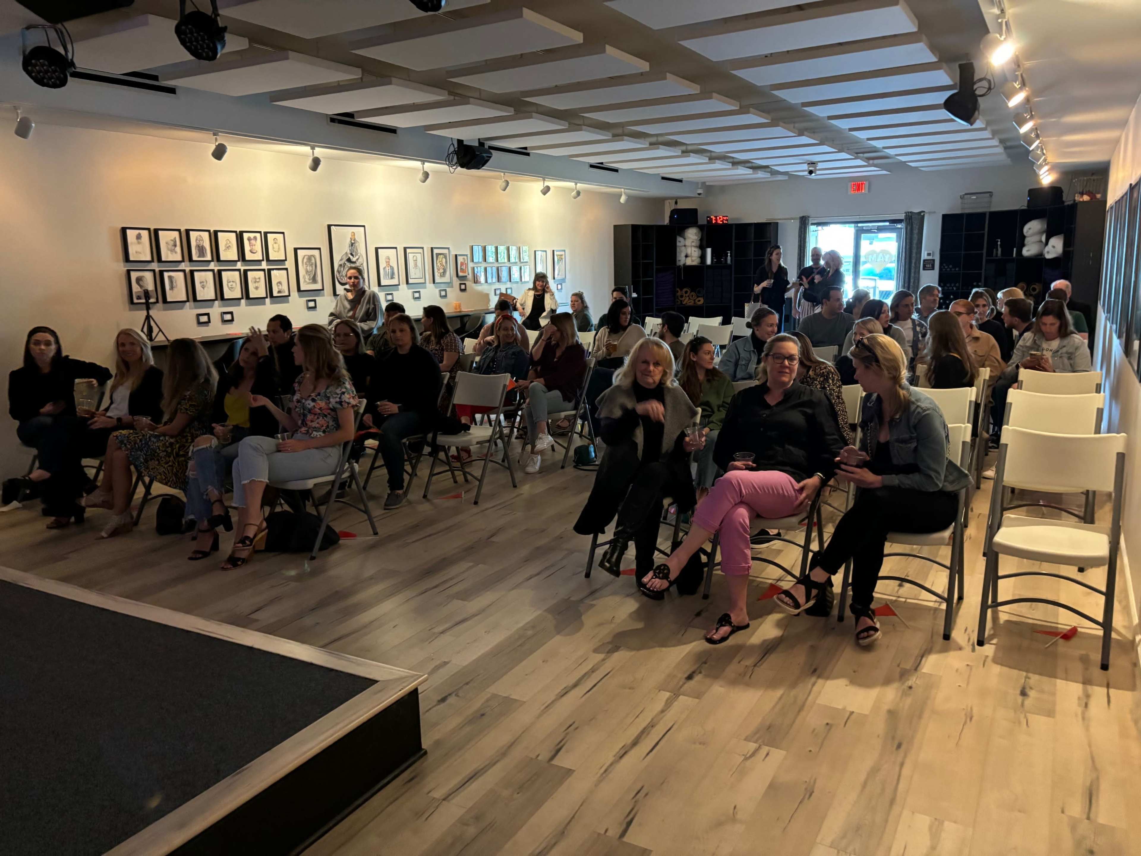 A large audience sits in a well-lit gallery space filled with framed artworks on the walls.