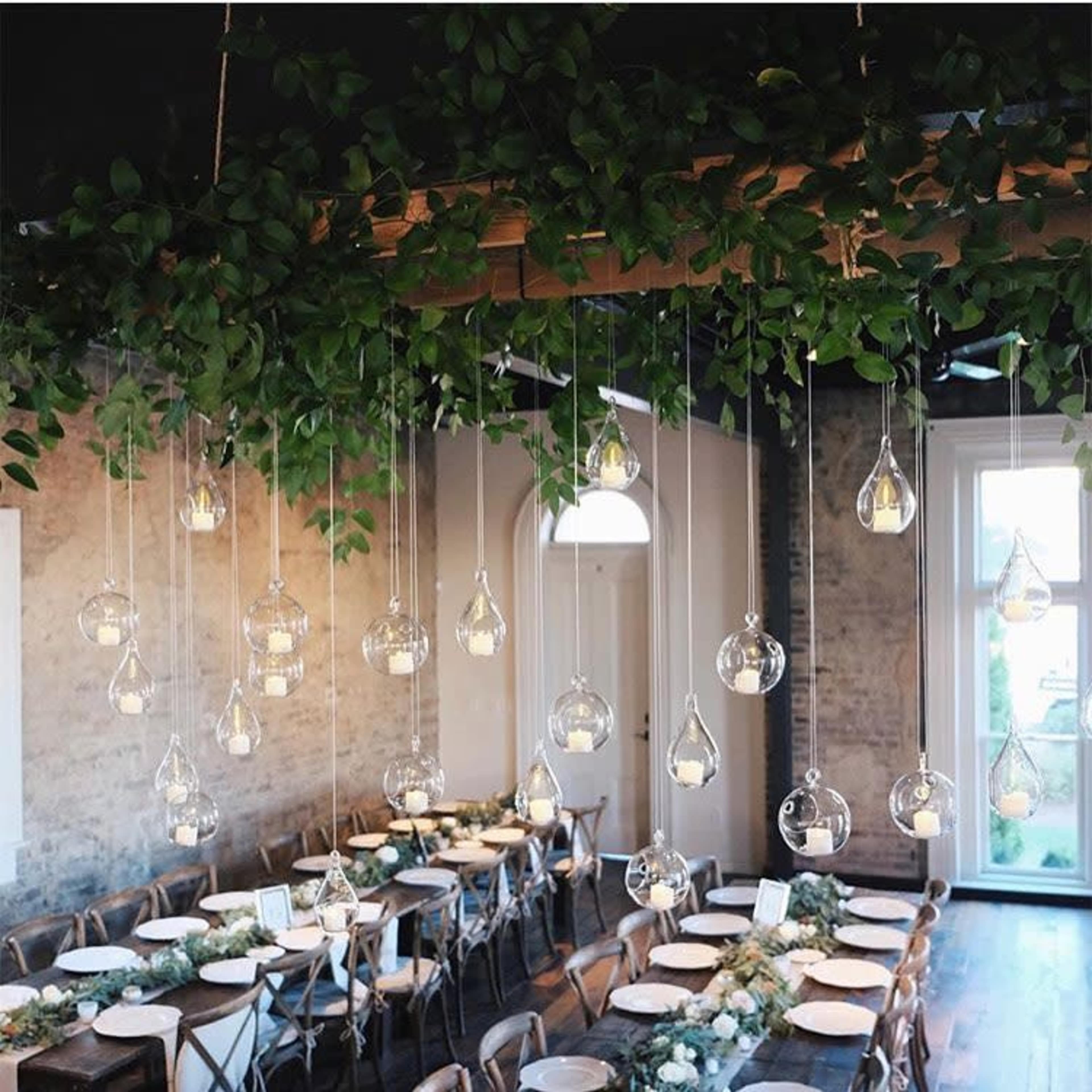 A decorated indoor dining area with hanging glass light fixtures and a long table set for a meal, surrounded by green foliage.