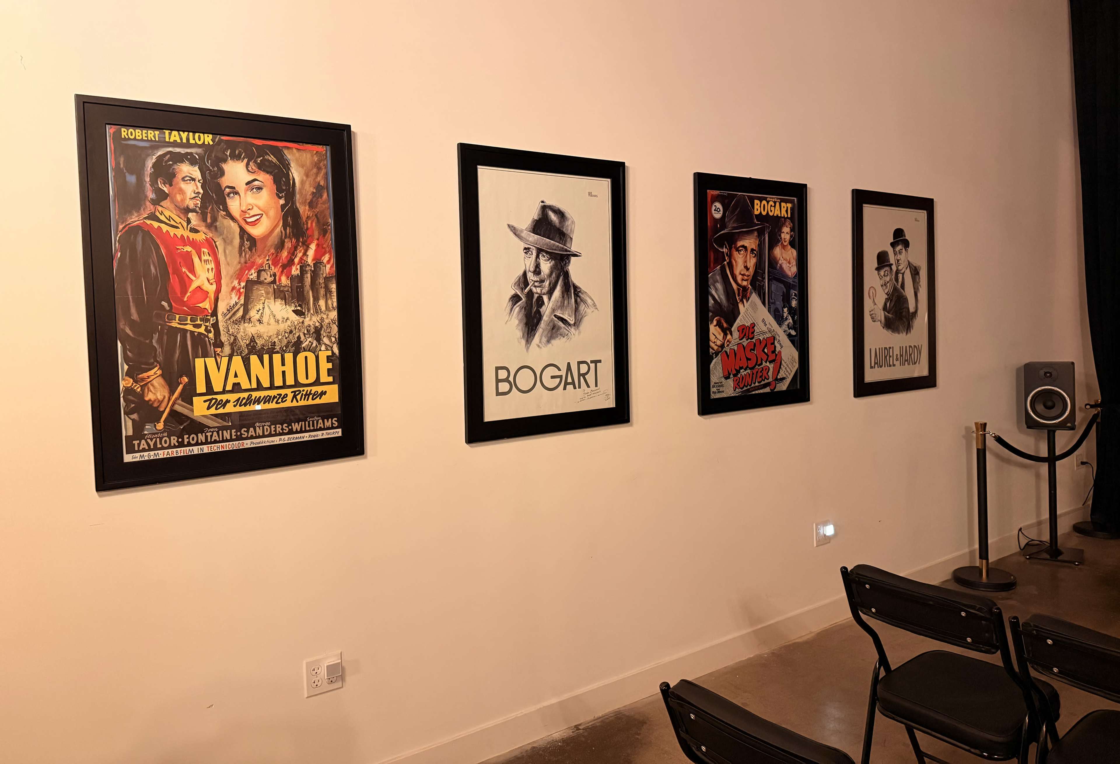 The image shows a wall with four framed movie posters, including titles such as "Ivanhoe" and "Bogart," alongside a speaker setup and chairs arranged for viewing.