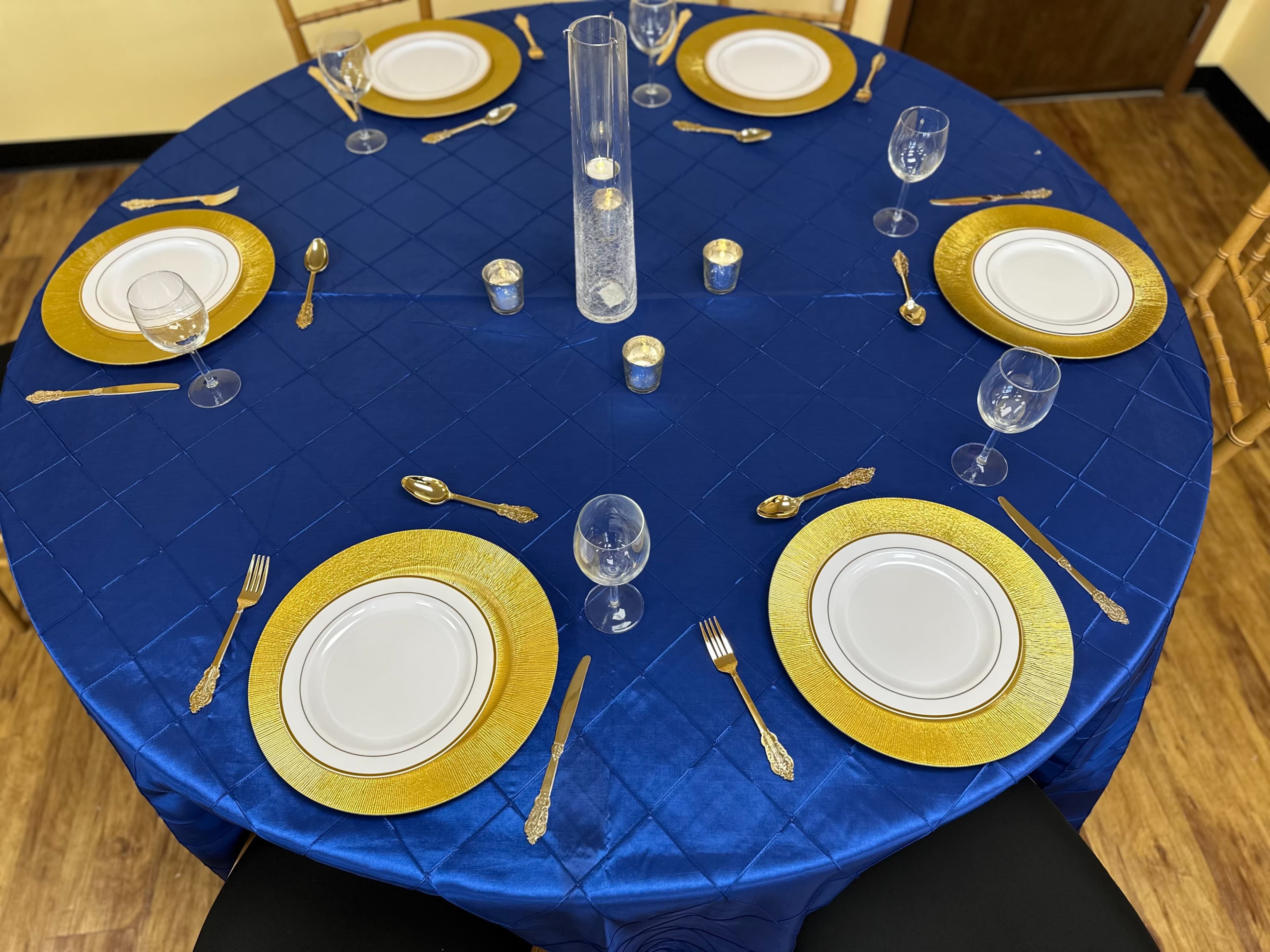 A circular table is set with gold-rimmed plates, silver utensils, clear glassware, and a tall centerpiece on a blue cloth.