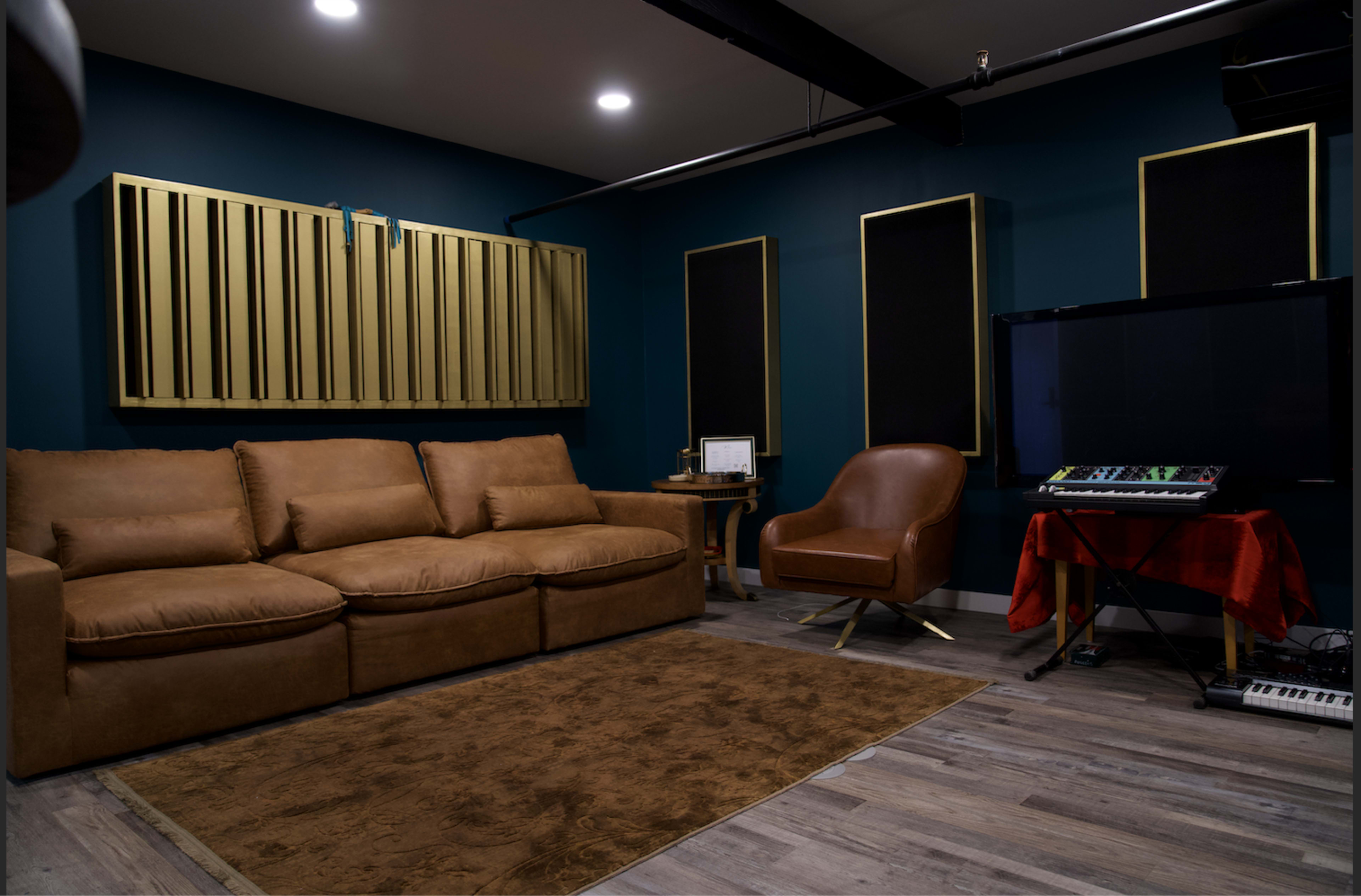 A soundproofed studio room featuring a brown sectional sofa, a wood chair, acoustic panels on the walls, and electronic equipment on a table.