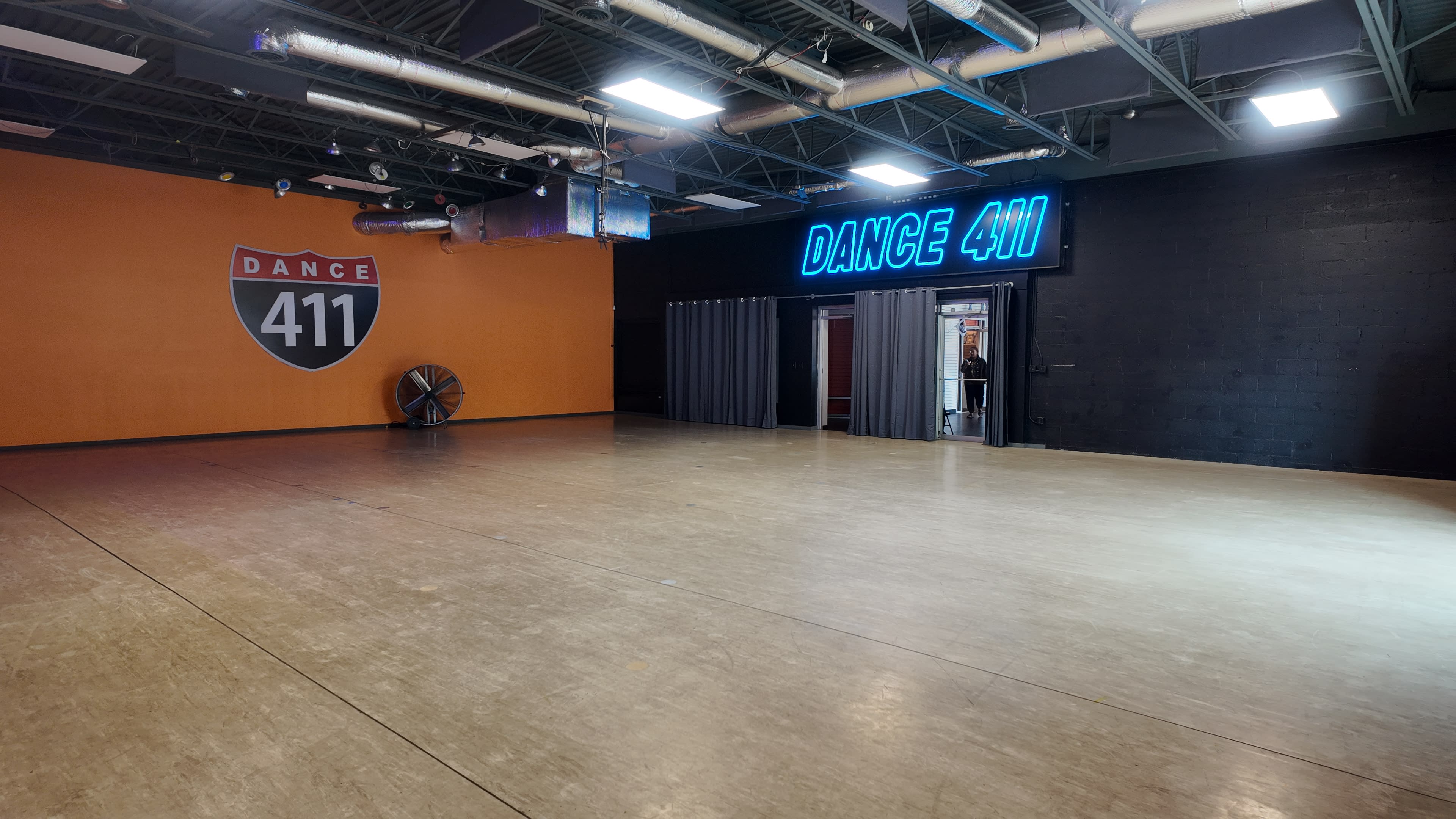 The image depicts a spacious dance studio with a bright orange wall featuring the logo "Dance 411" and a large neon sign that spells out "DANCE 411."