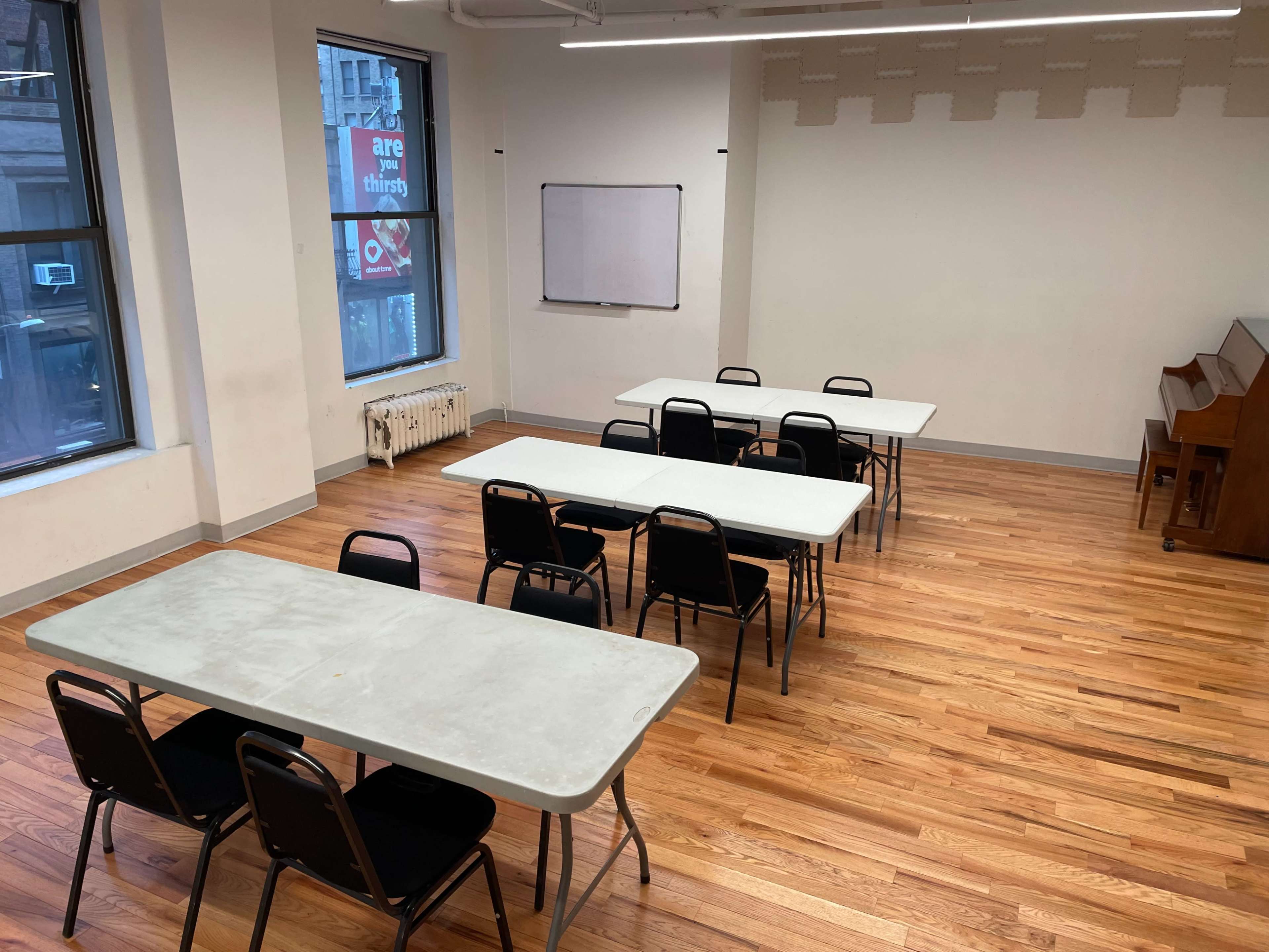 A spacious room with wooden flooring, featuring several set up tables and chairs, a whiteboard on the wall, and a piano in the corner.