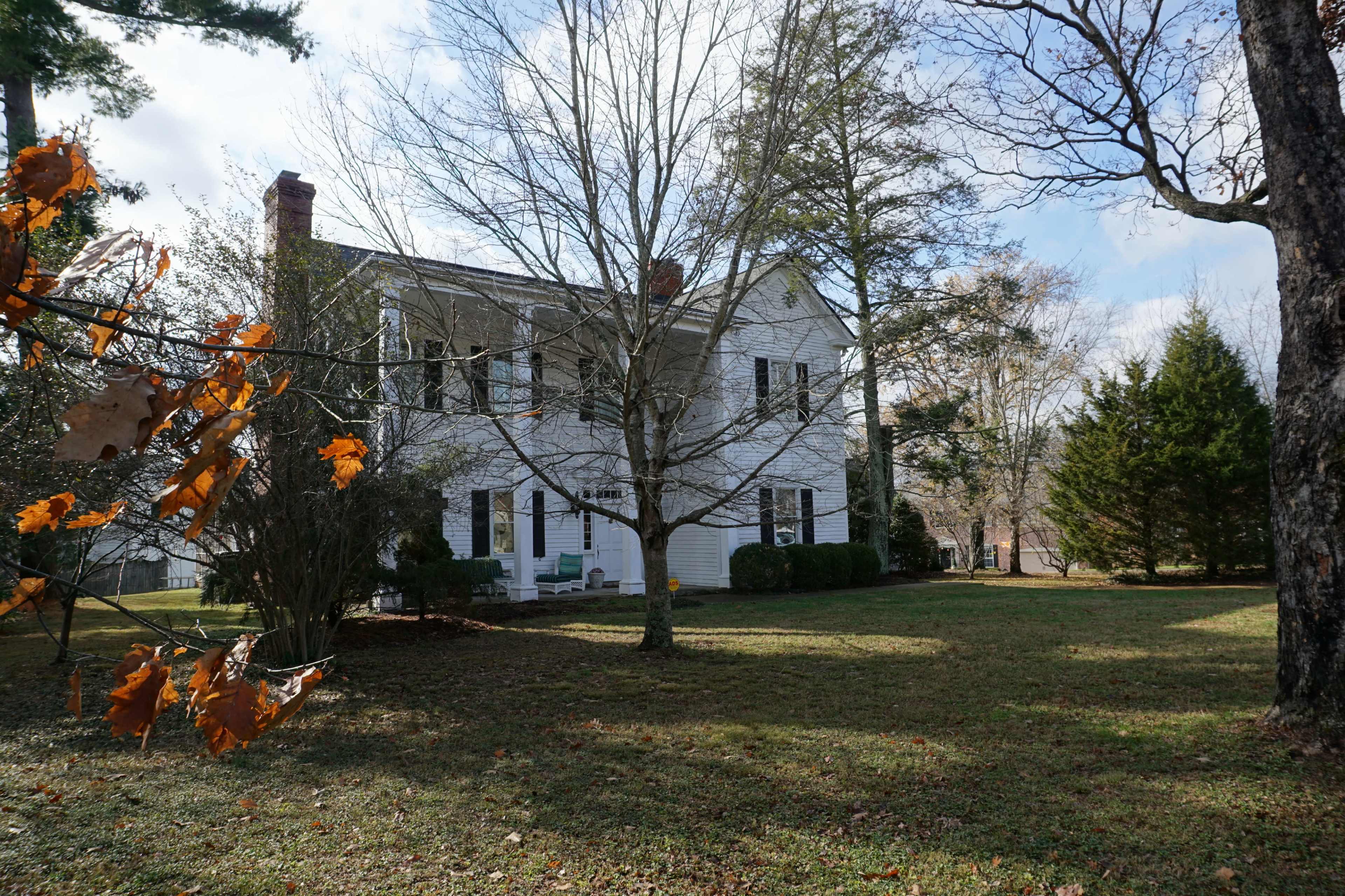 A two-story white house stands among trees on a grassy lawn, with autumn leaves scattered on the ground.