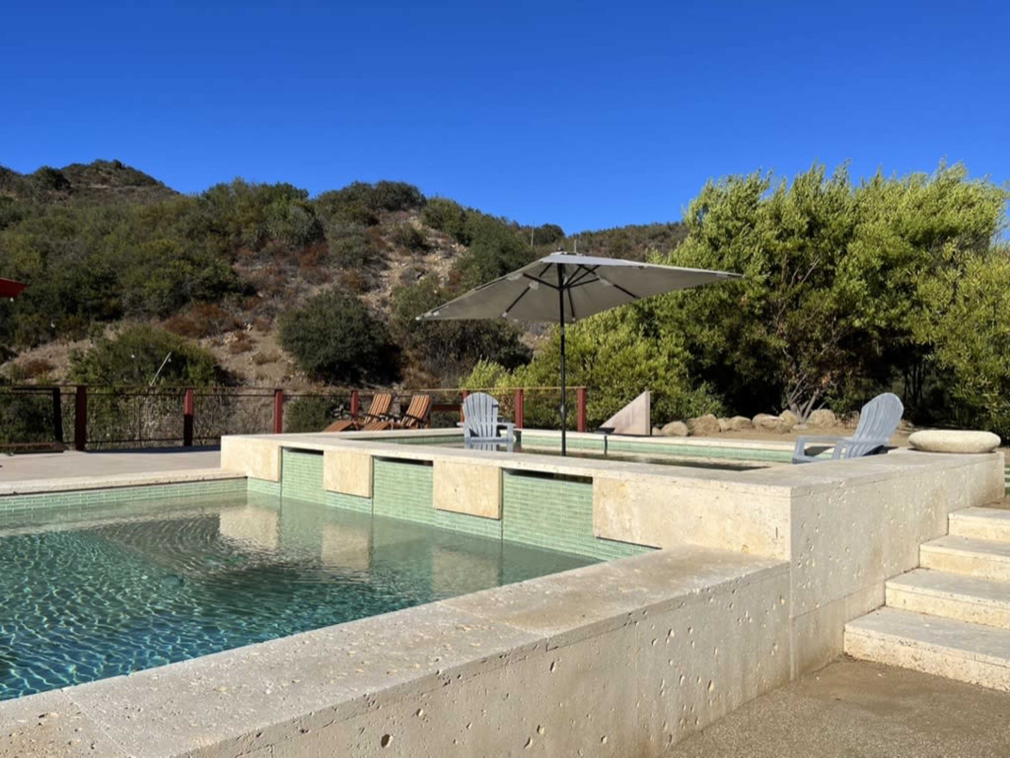 A swimming pool with a sun umbrella and lounge chairs is surrounded by greenery and hills.