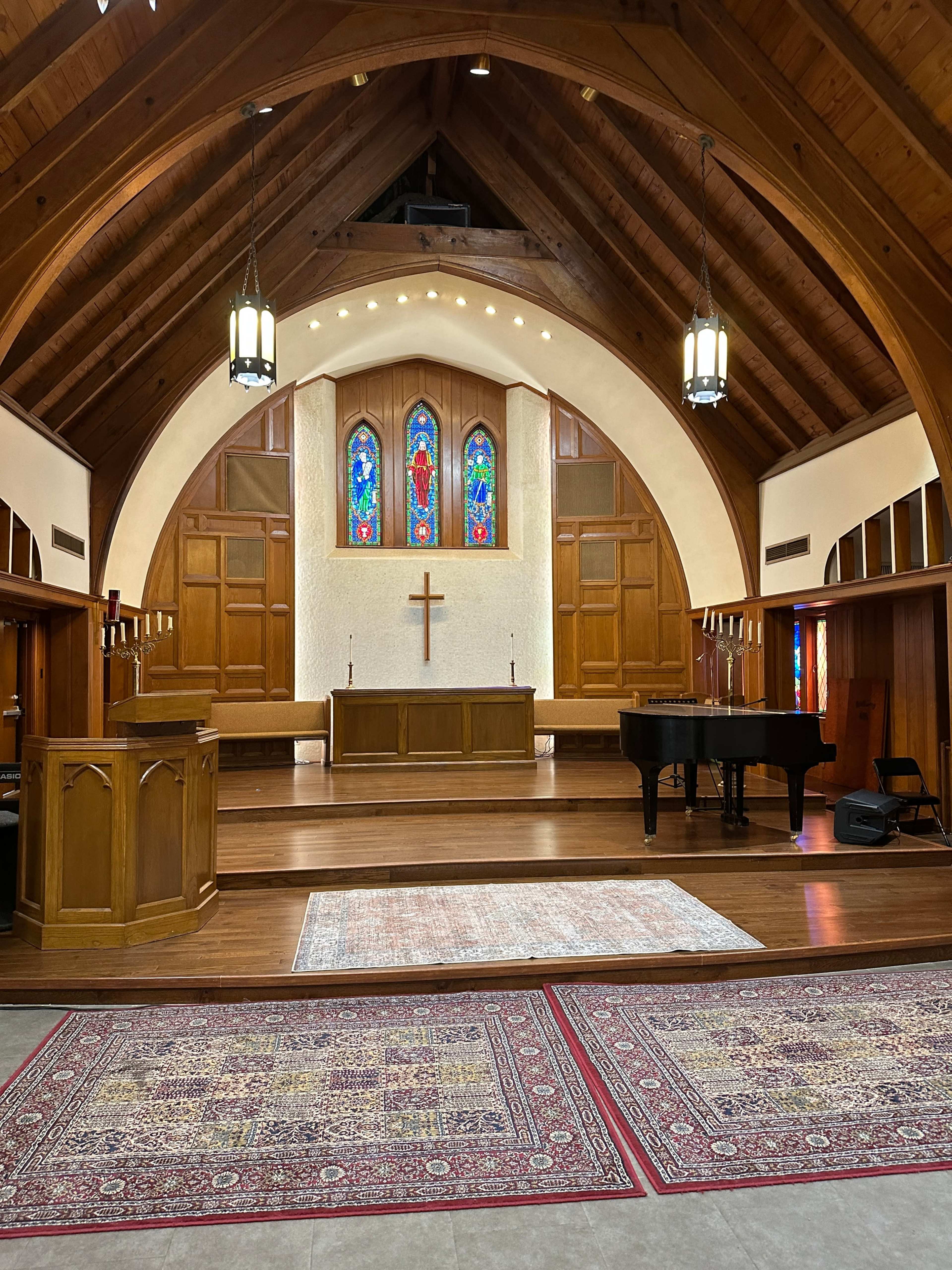 The interior of a church features a wooden arch ceiling, stained glass windows, a central cross, and a grand piano on a polished floor.