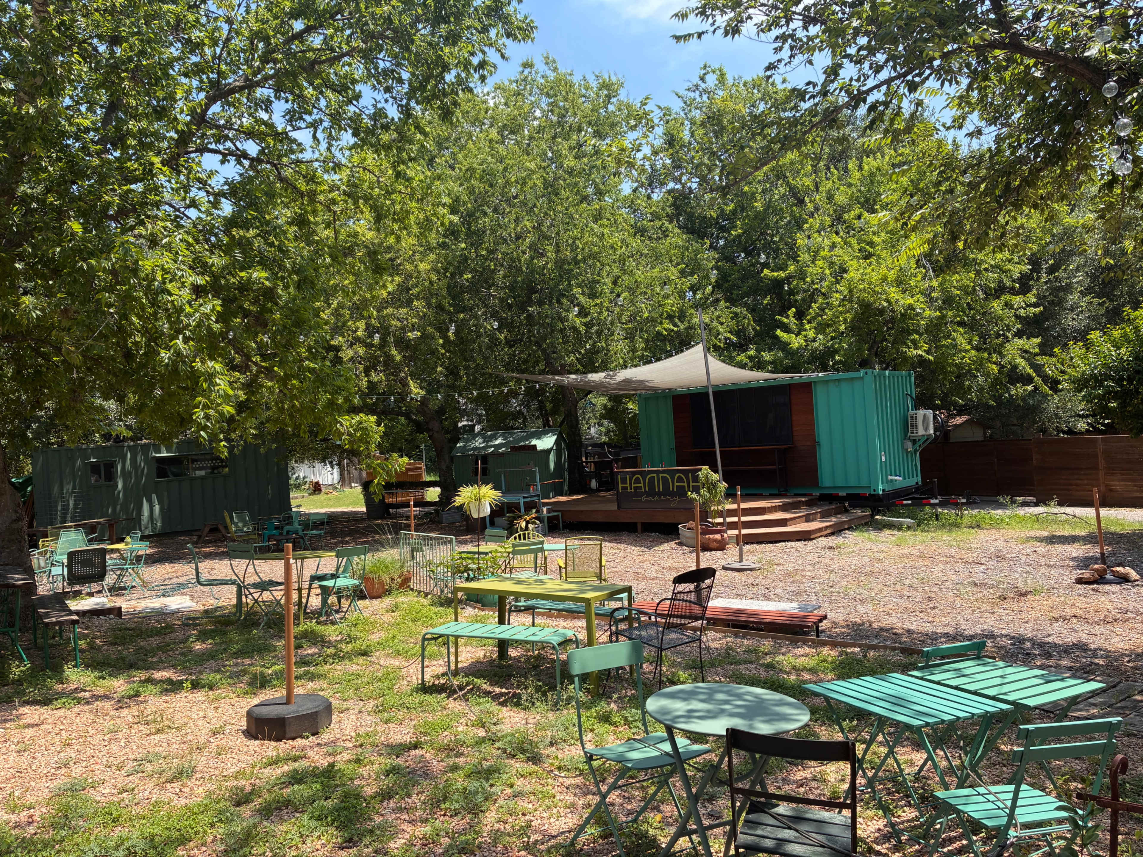 The image shows a landscaped outdoor area with green metal tables and chairs, surrounded by trees and featuring a small building in the background.