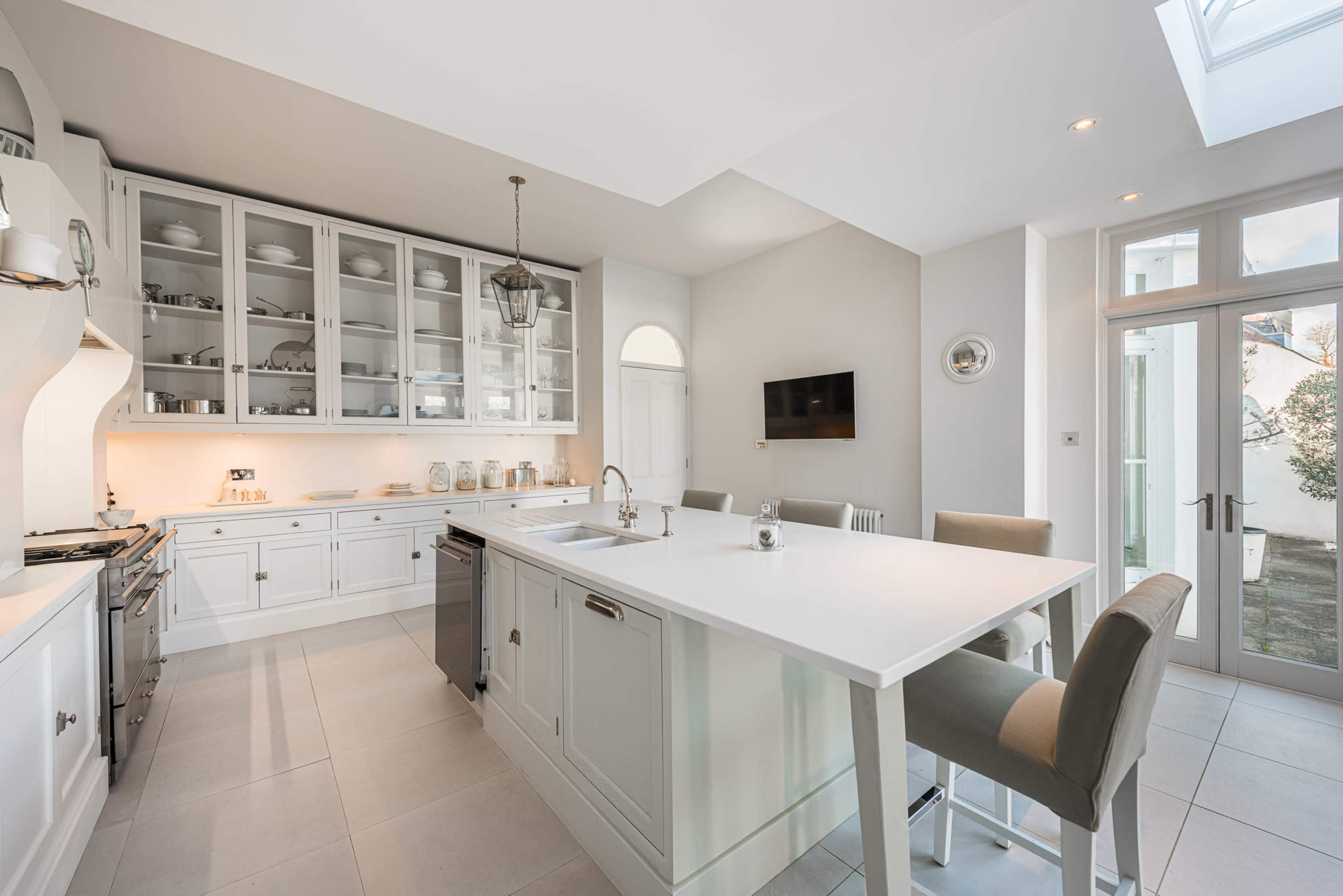 The image shows a modern kitchen with white cabinetry, a central island with bar seating, and large windows allowing natural light to enter the space.