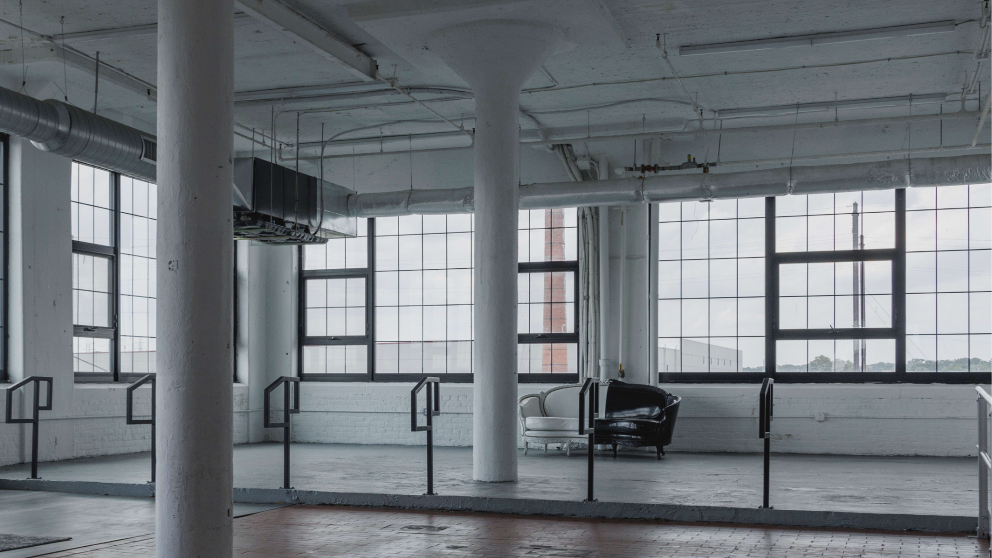 The image shows an empty industrial space with large windows, exposed pipes, and a black armchair near the wall.