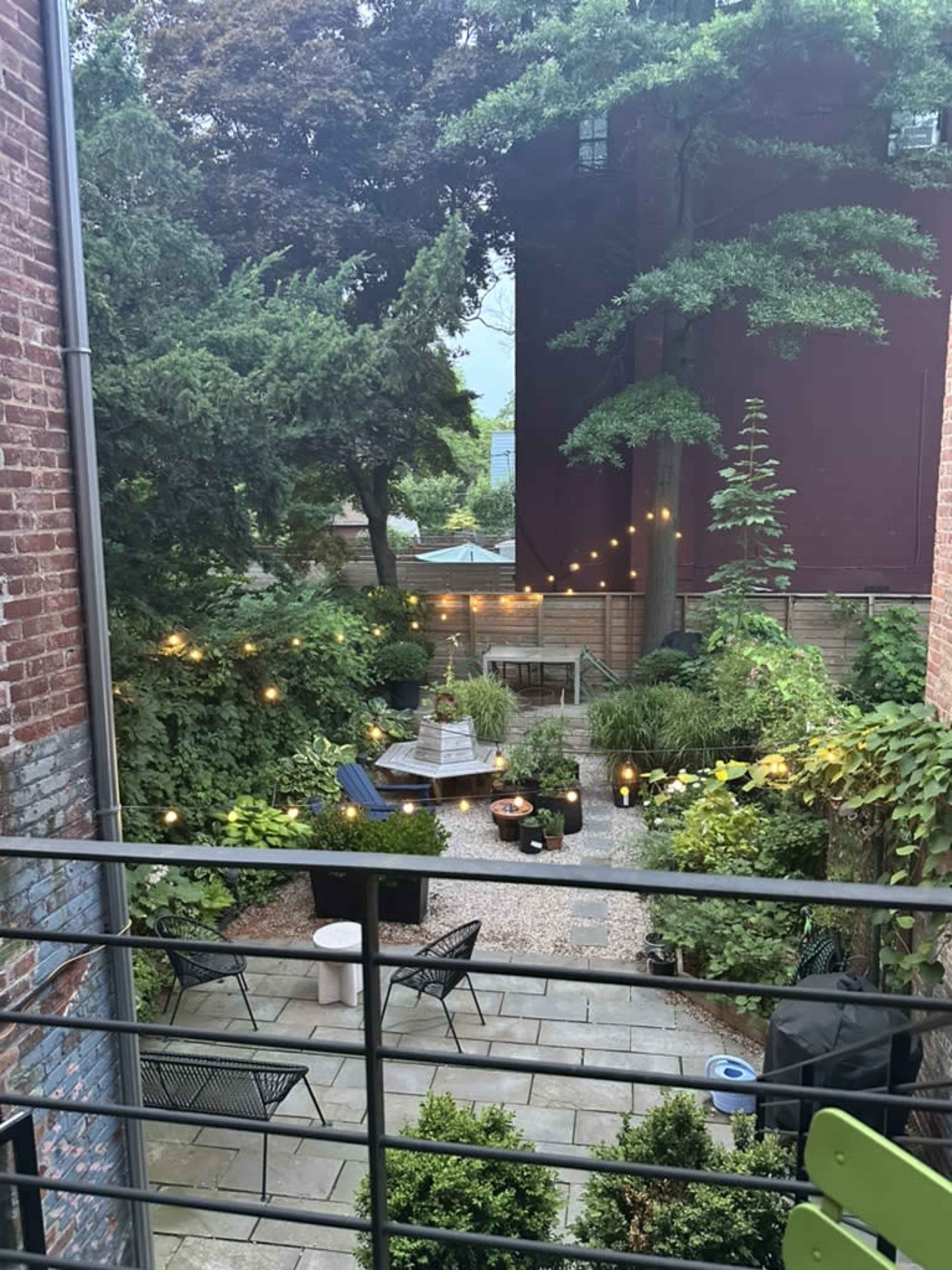 The image shows a backyard garden with stone pathways, seating areas, potted plants, and string lights illuminating the space.