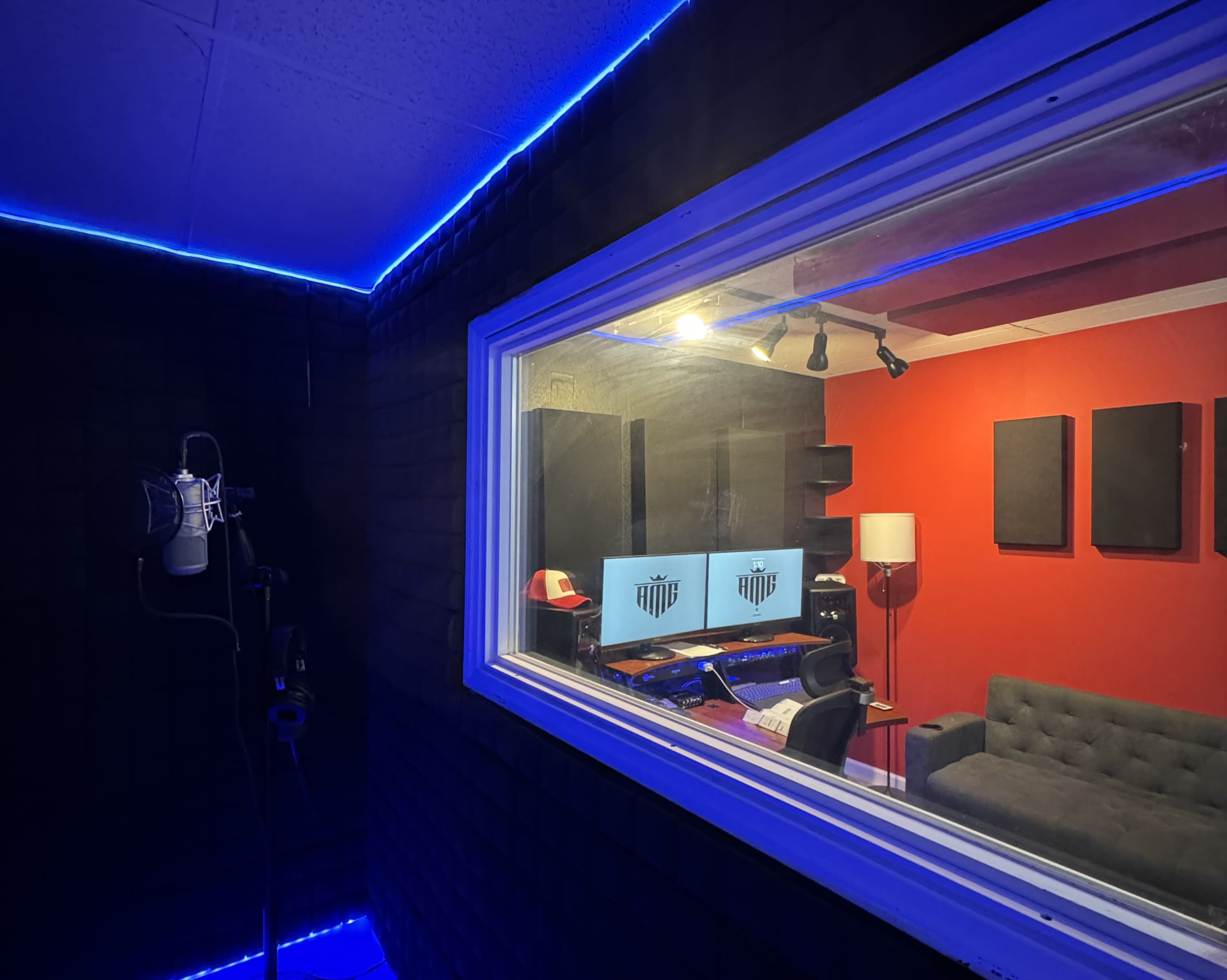 The image shows a soundproof recording studio with a microphone and monitors visible through a glass window, featuring a red wall and a couch.