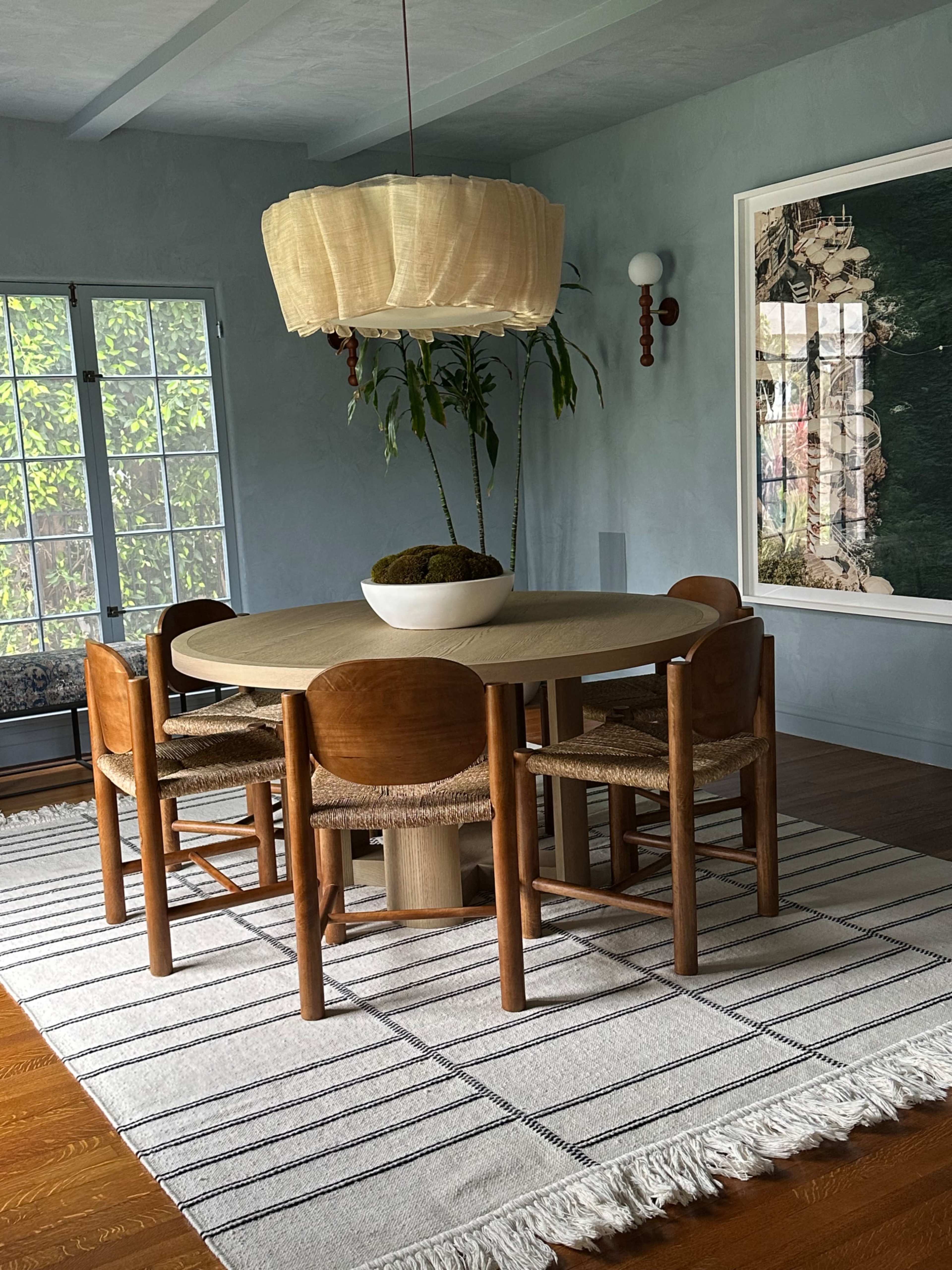 A round wooden dining table with six chairs is placed on a striped rug beneath a large pendant light in a softly lit room with blue walls and a large artwork.