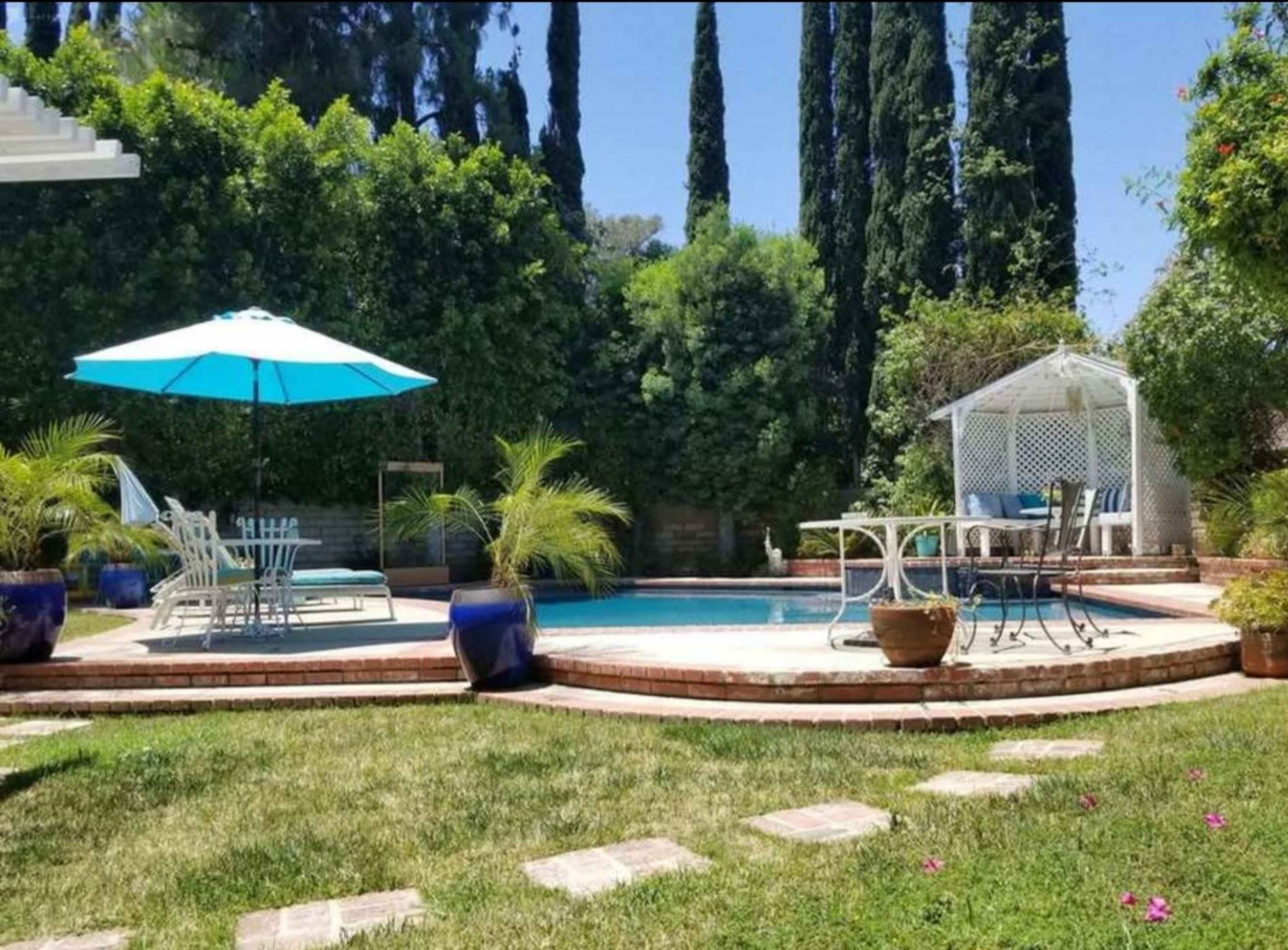 A backyard features a swimming pool surrounded by stone pathways, lounge furniture, and tall cypress trees, with a blue umbrella providing shade.