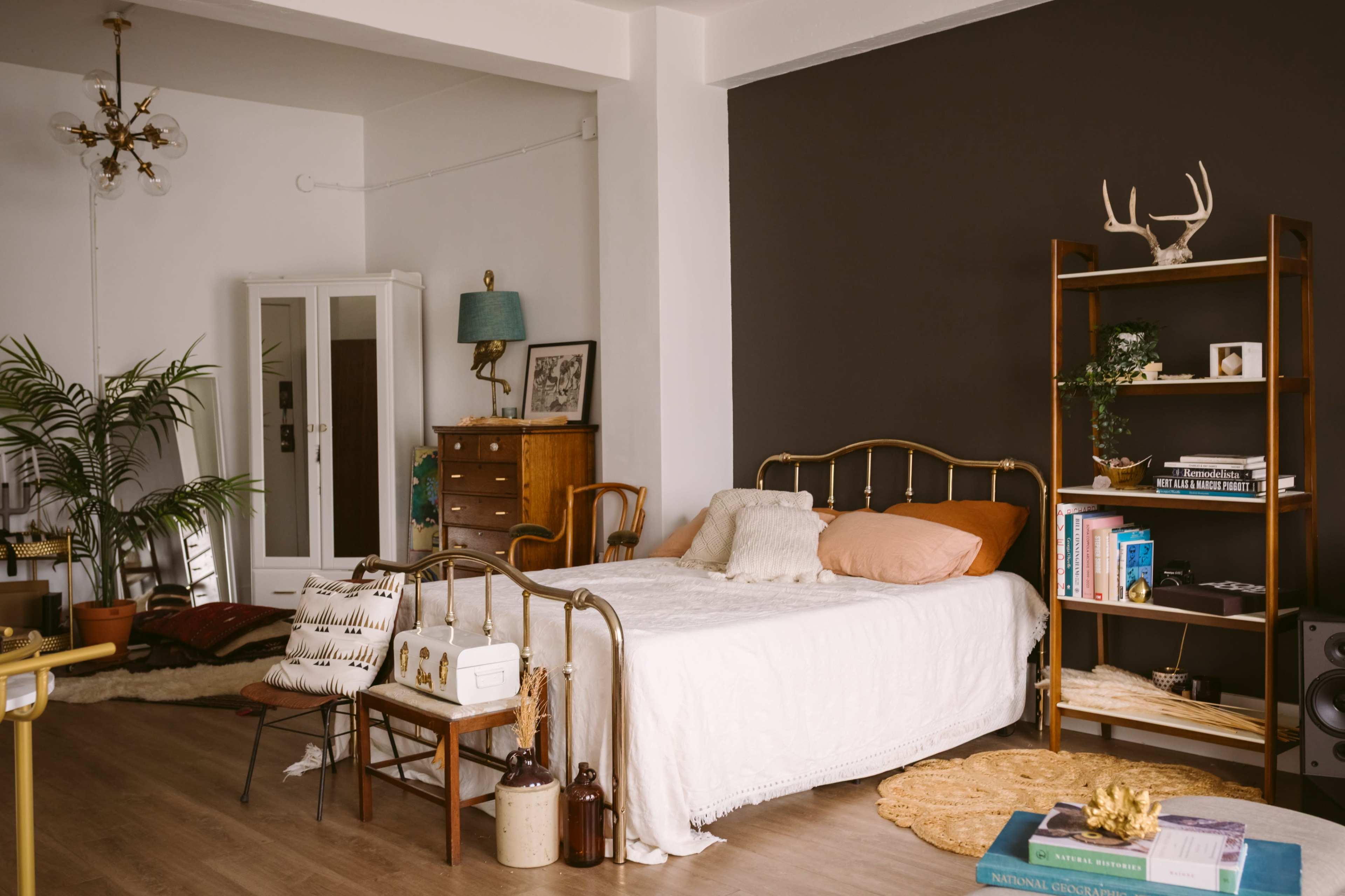The scene shows a cozy bedroom featuring a brass bed with a white bedspread, a wooden dresser, a bookshelf, and various decorative elements against a dark accent wall.