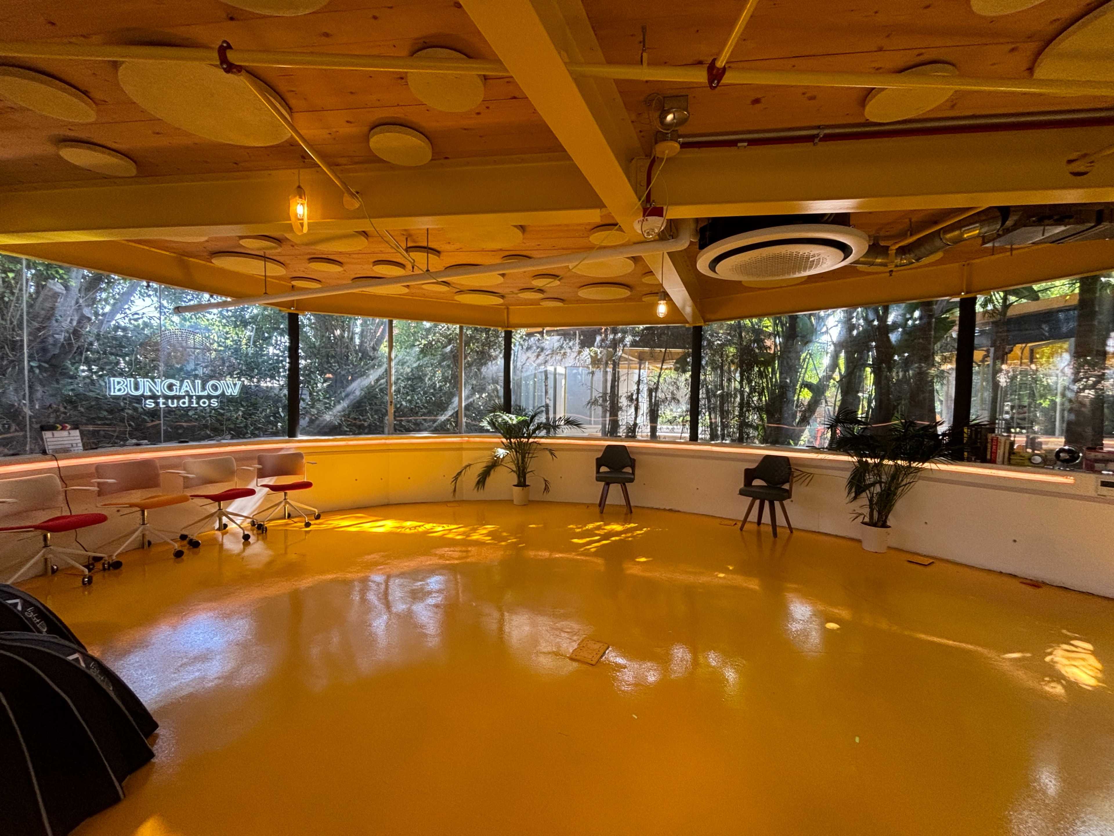 The image shows a spacious interior of a studio with a yellow floor, large windows surrounded by greenery, and modern furniture including chairs and workstations.