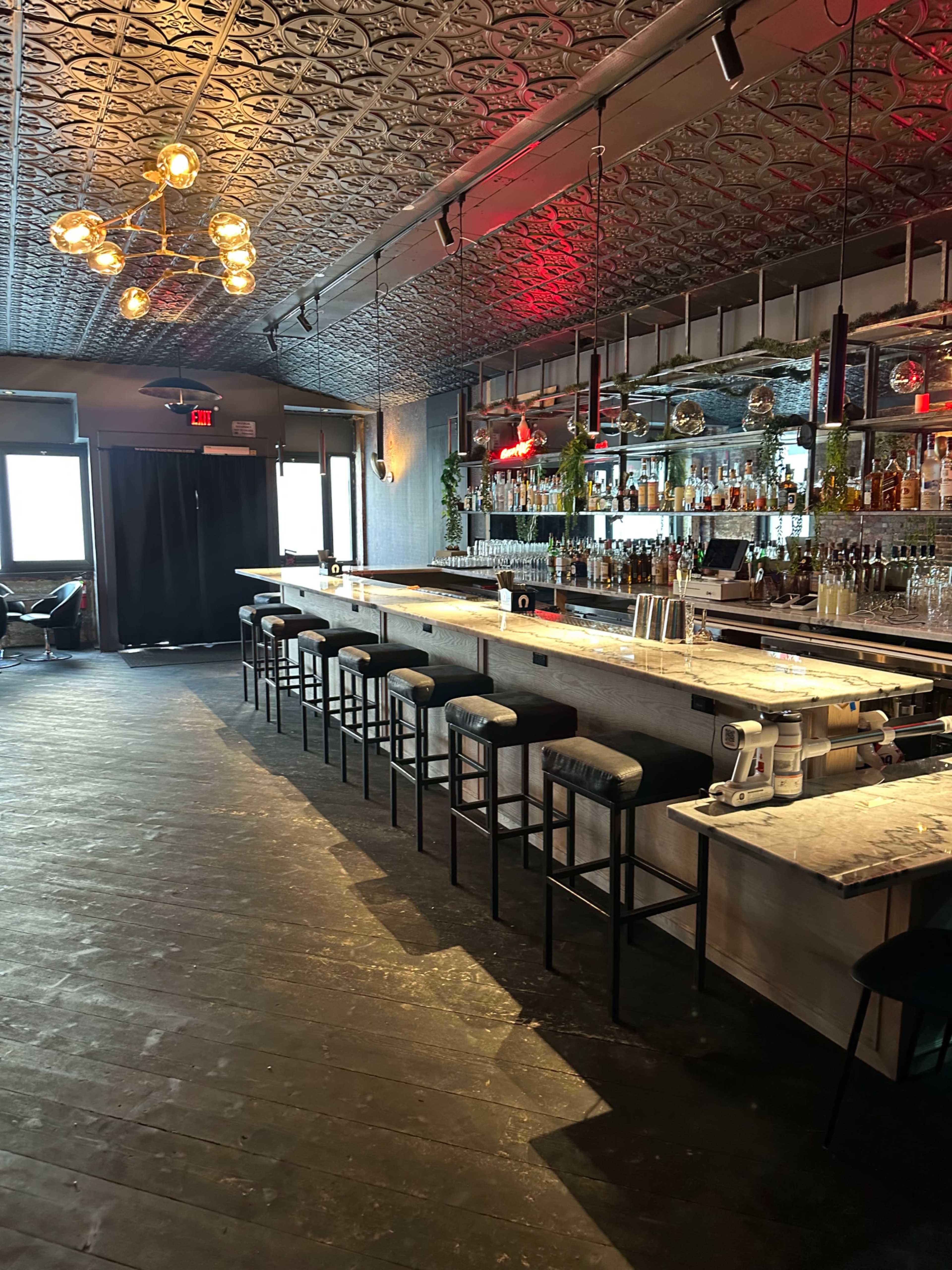 The image shows a modern bar interior featuring a long marble countertop, high stools, and a well-stocked back bar with shelves of liquor and decorative plants.