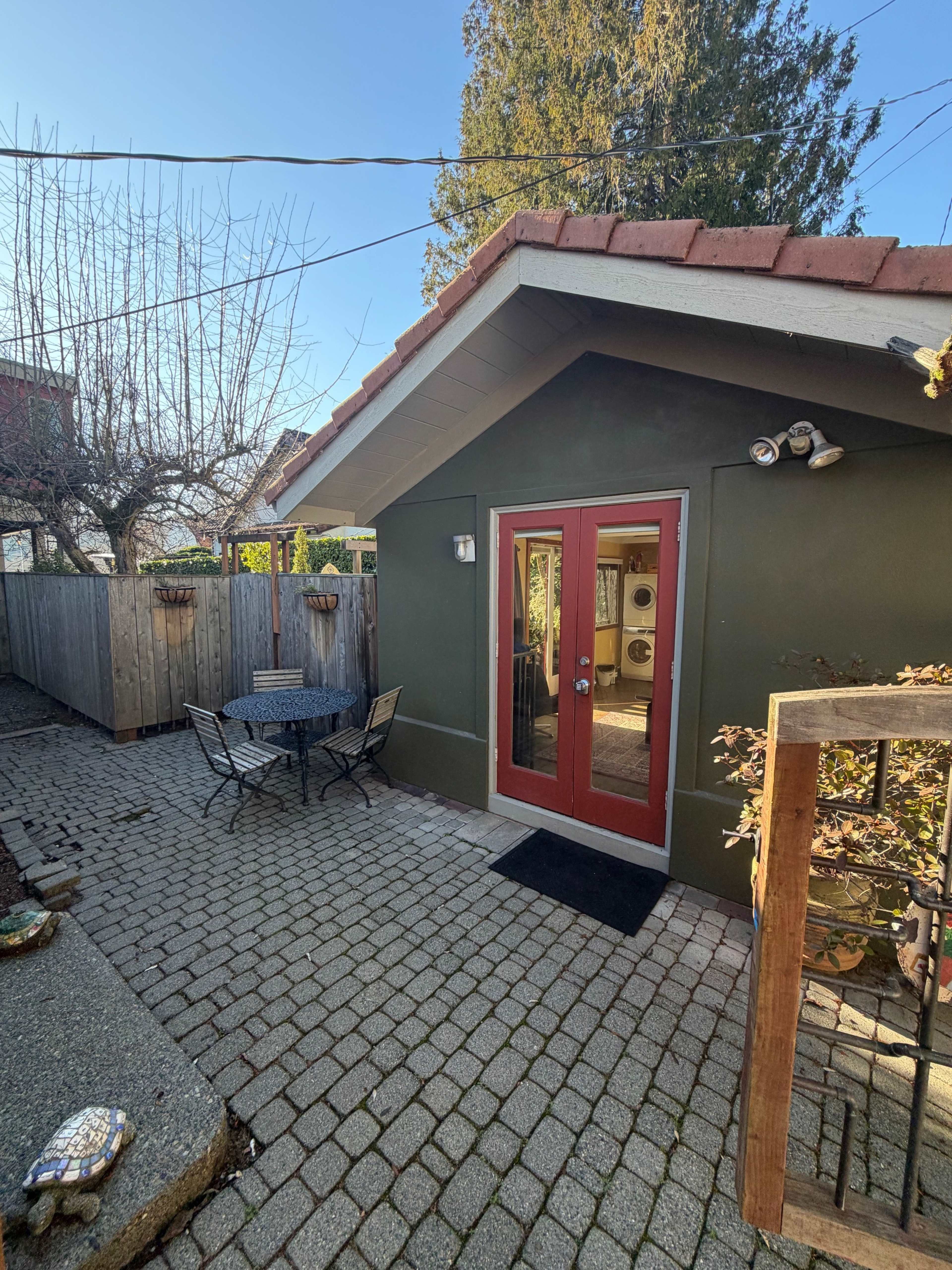 The image shows a small outdoor patio with a stone-paved area, featuring a round table and chairs in front of a green shed-like structure with red double doors.