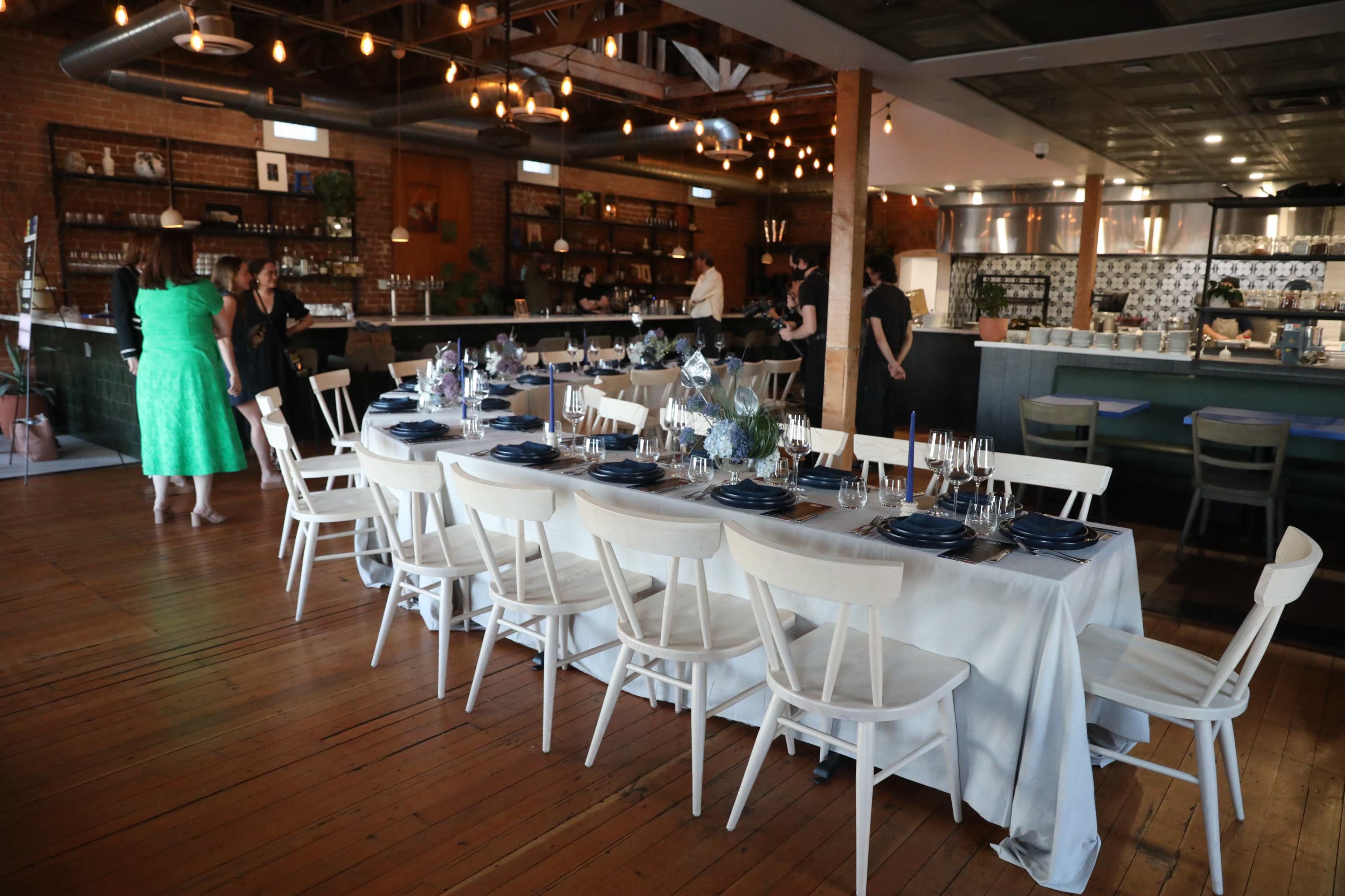A long table is set with blue plates and silverware in a modern restaurant with exposed brick walls and overhead string lights.