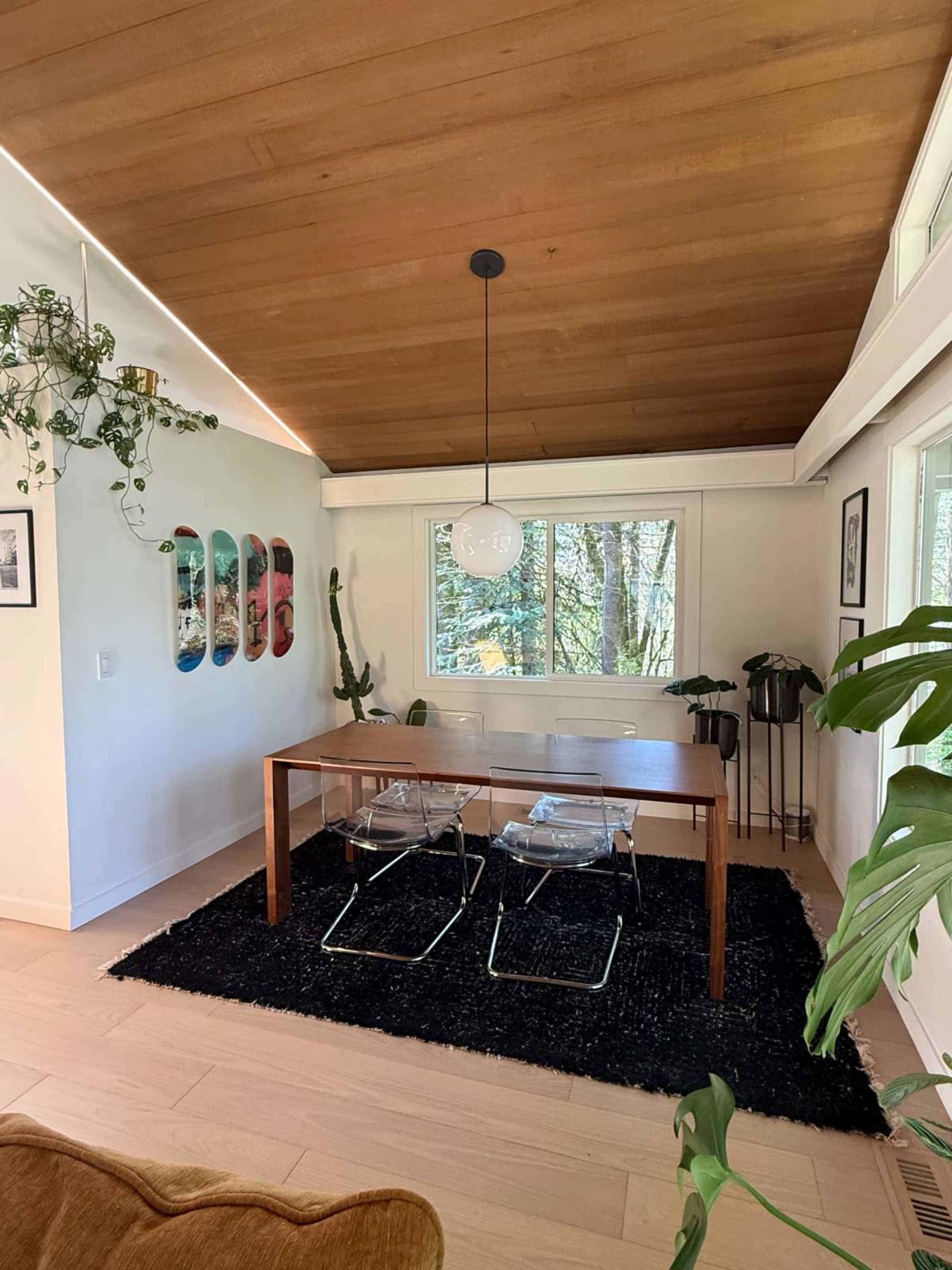 A dining area features a wooden table with two chairs, surrounded by plants and illuminated by a pendant light.