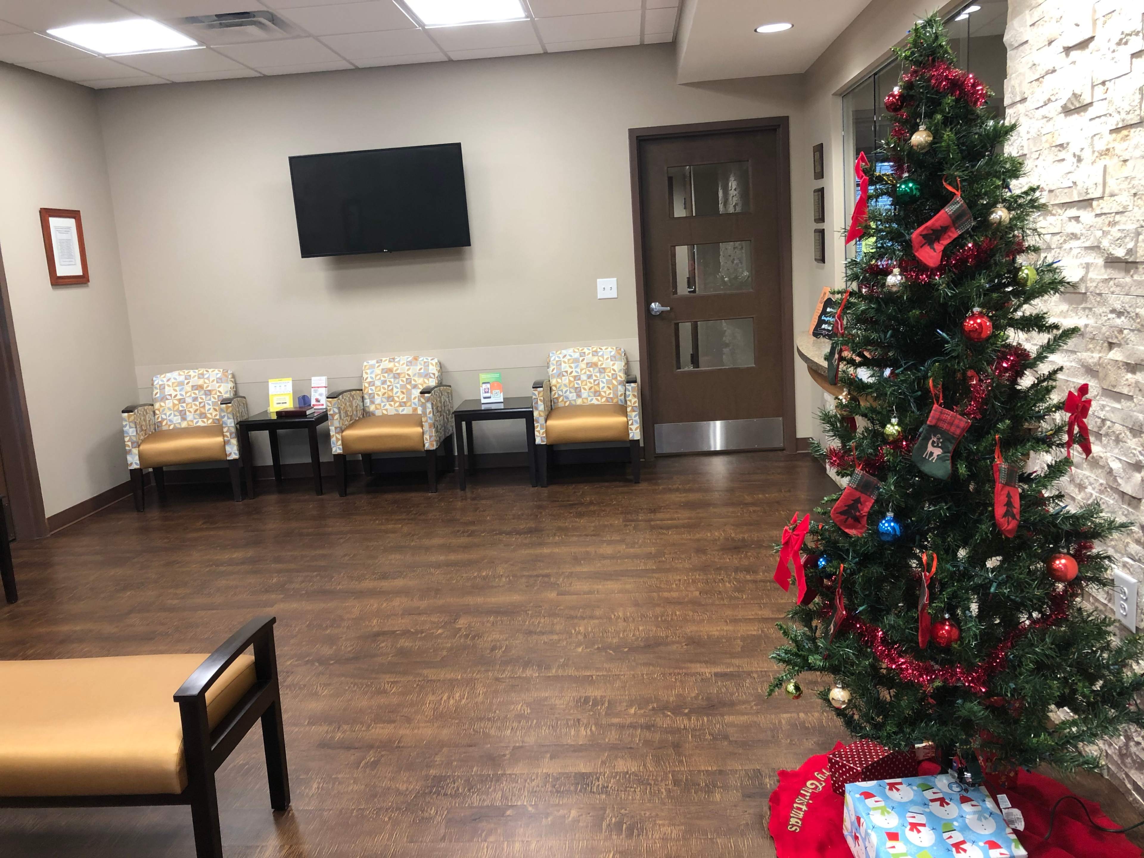 A waiting room with three patterned chairs, a flat-screen television on the wall, and a decorated Christmas tree in the corner.