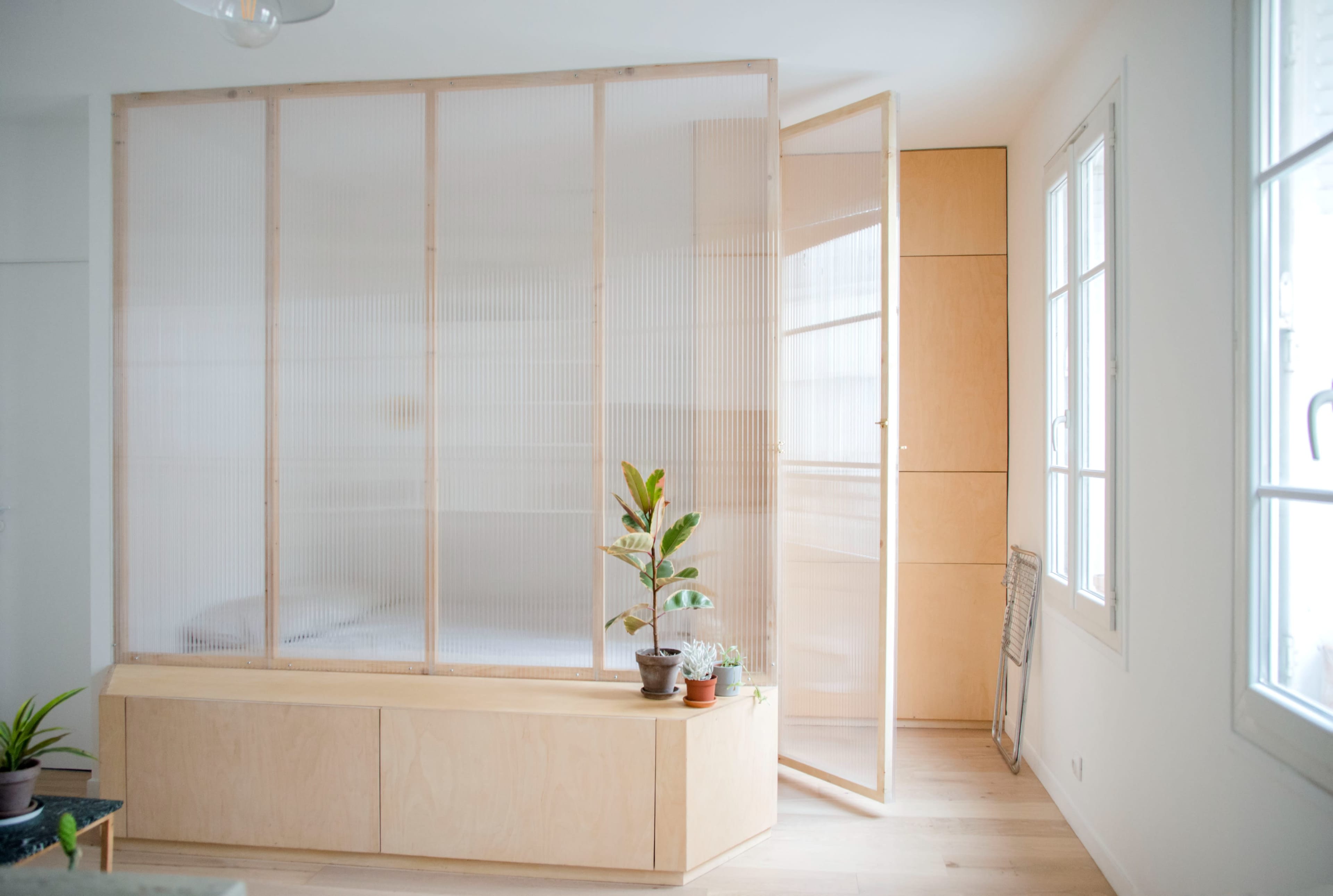 The image shows a lightly furnished room with a wooden partition separating a sleeping area, featuring a few potted plants in the foreground.