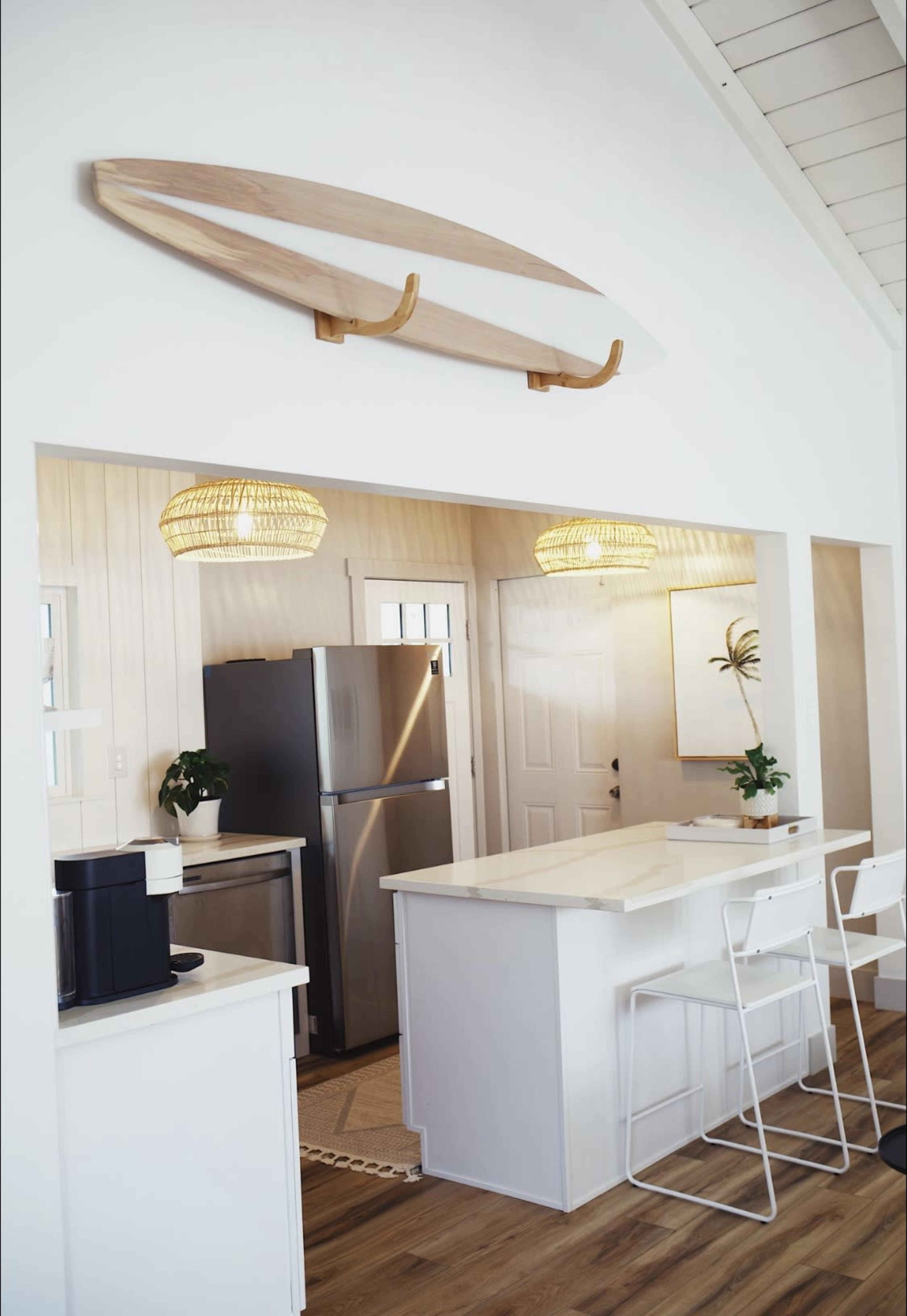 A modern kitchen with a white island, stainless steel appliances, and a surfboard mounted on the wall above.