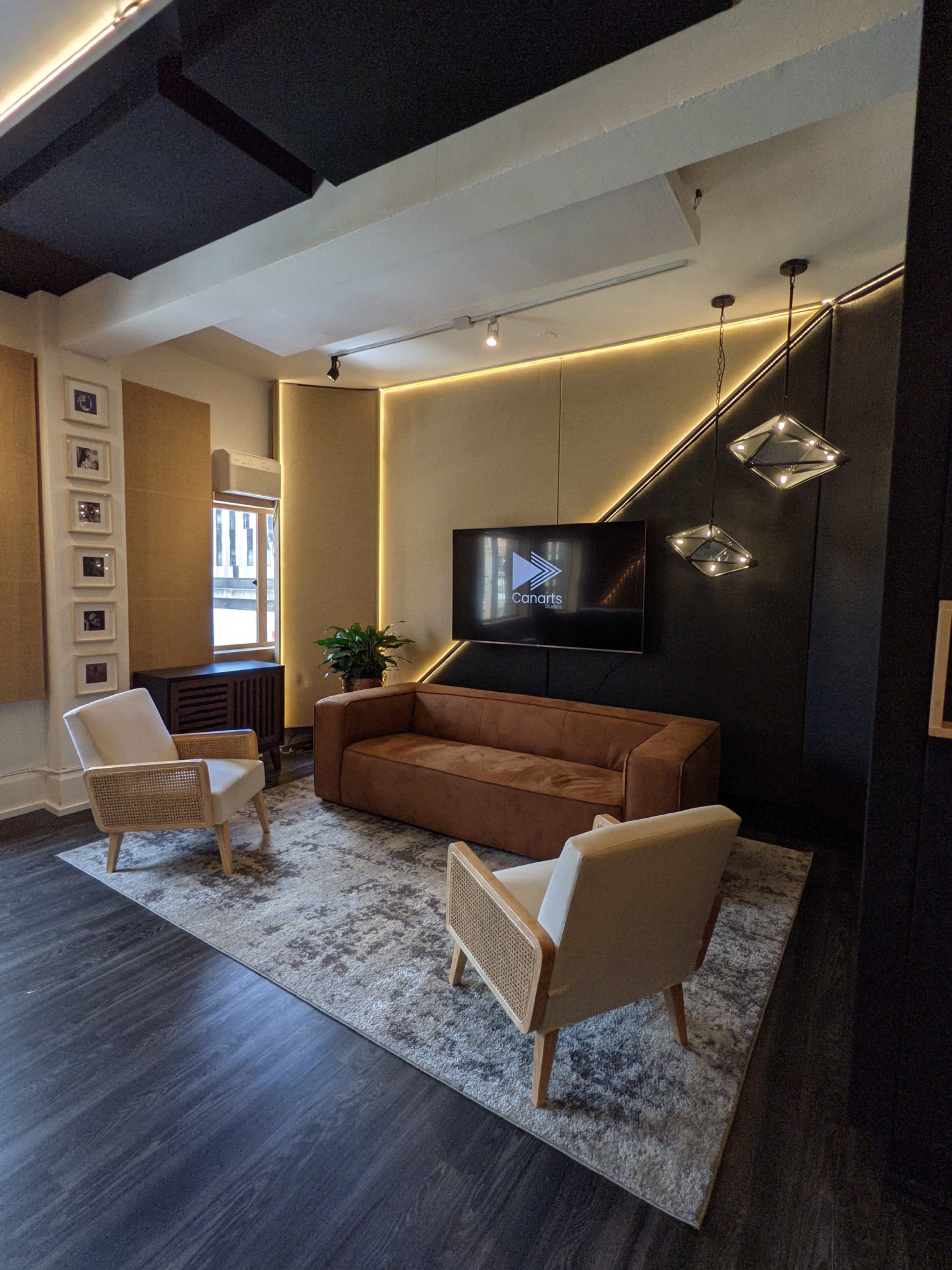 A modern living space featuring a brown couch, two white armchairs, a large television mounted on a black wall, and decorative lighting overhead.