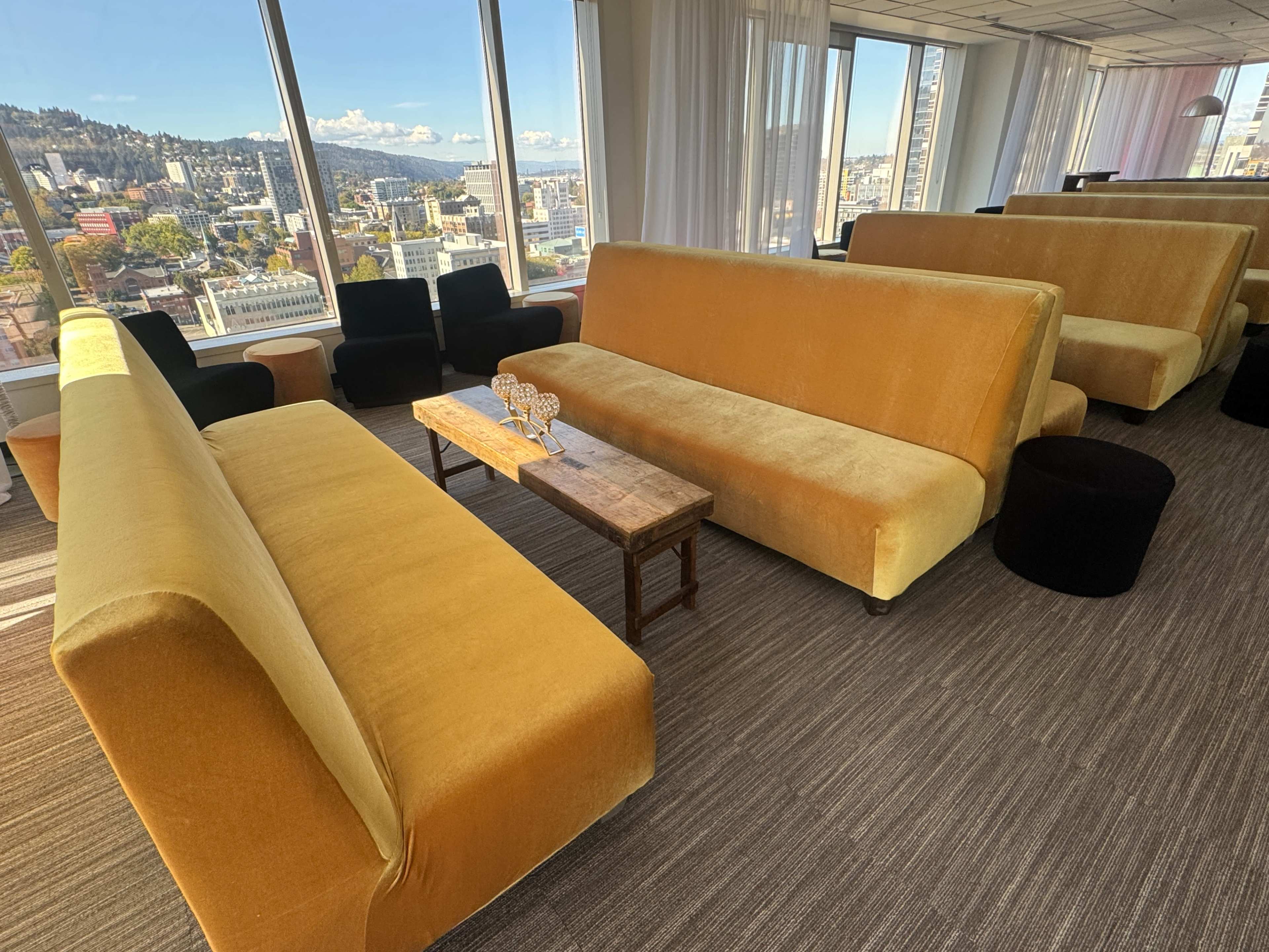The image shows a modern, spacious lounge area with yellow and black seating, a wooden coffee table, and large windows overlooking a cityscape.
