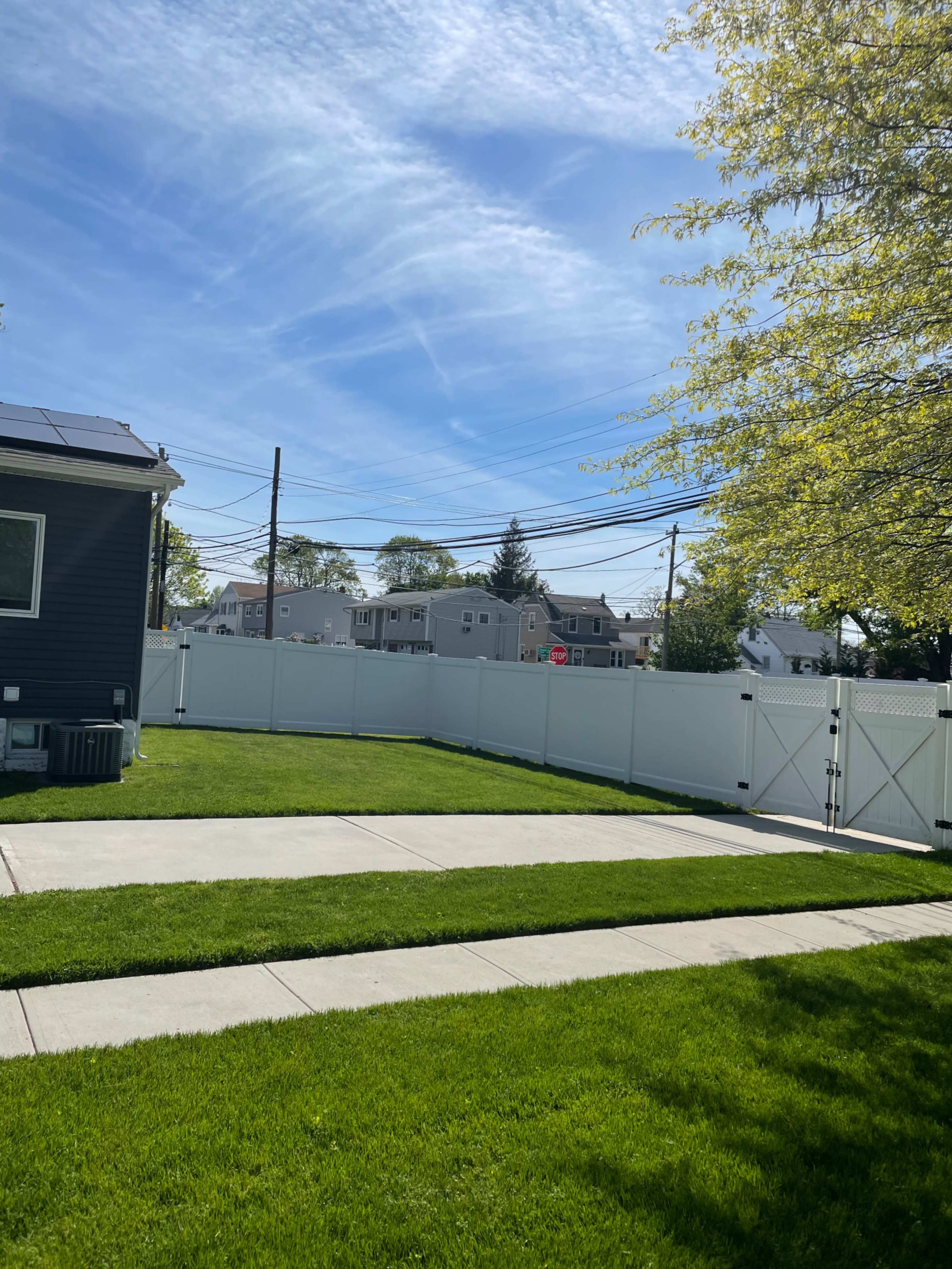 A landscaped yard features a clear blue sky, a white fence, and a paved walkway leading to a house.