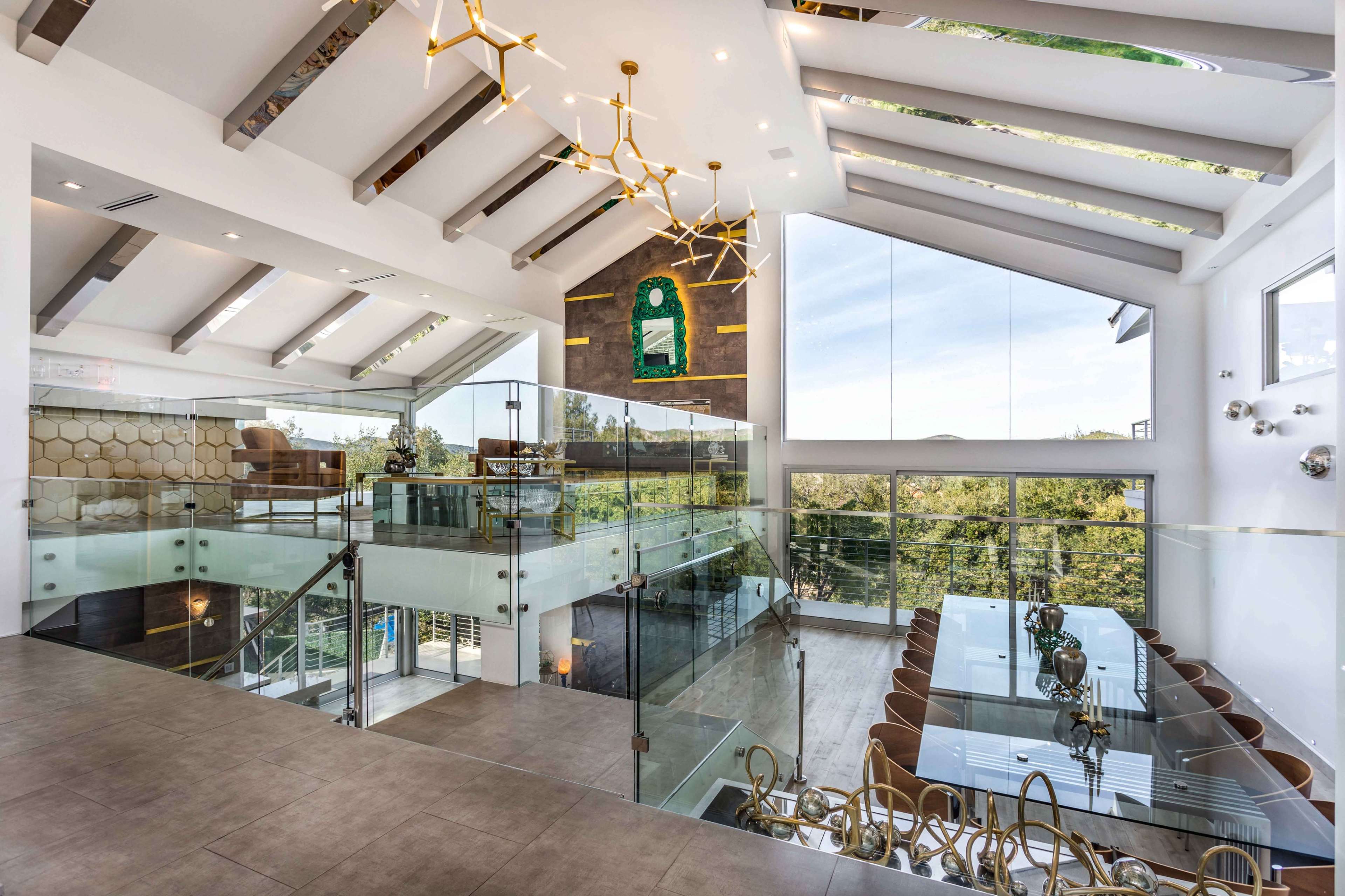 A spacious, modern interior features a glass dining table surrounded by elegant chairs, with a view of greenery through large windows and a staircase with a glass railing.