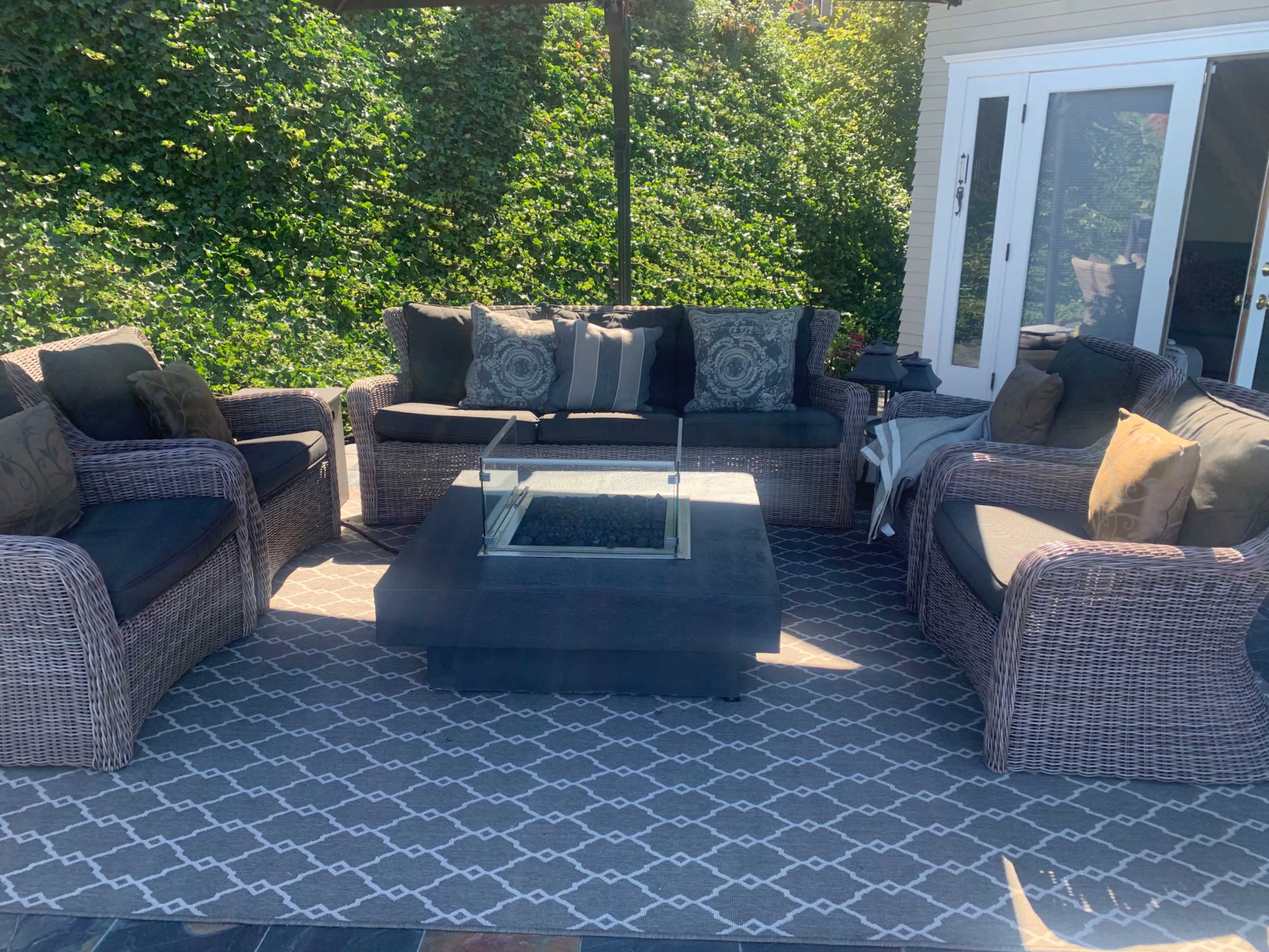 A covered outdoor seating area features a wicker sofa set with cushions, a glass-top coffee table, and a patterned rug.