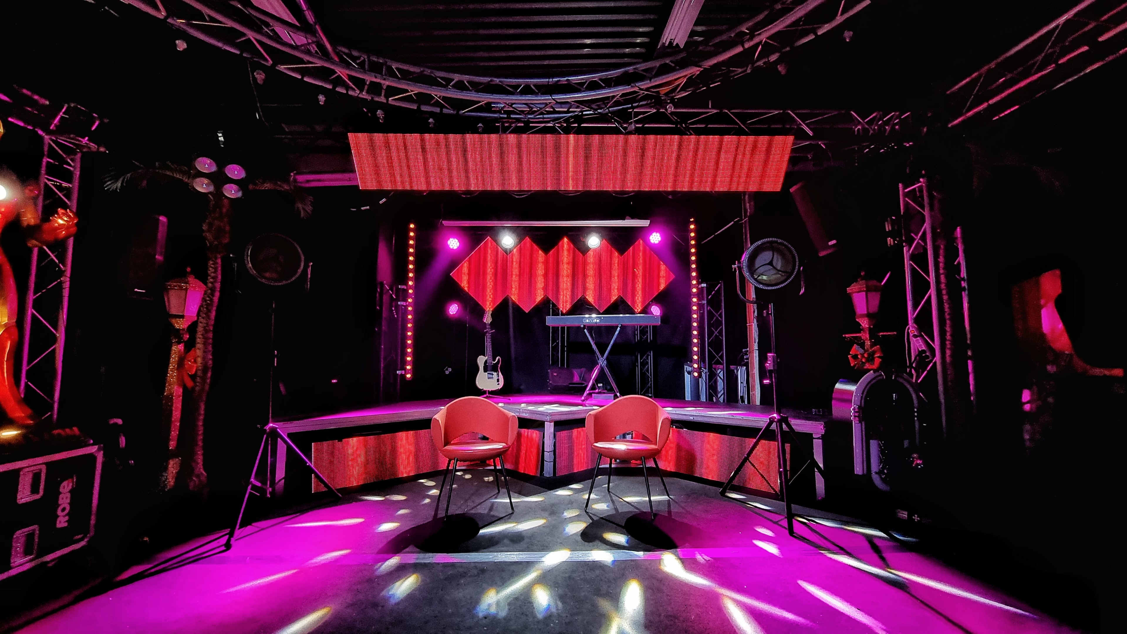 A stage setup featuring two chairs, a guitar, and a keyboard under vibrant stage lights and a decorative backdrop.