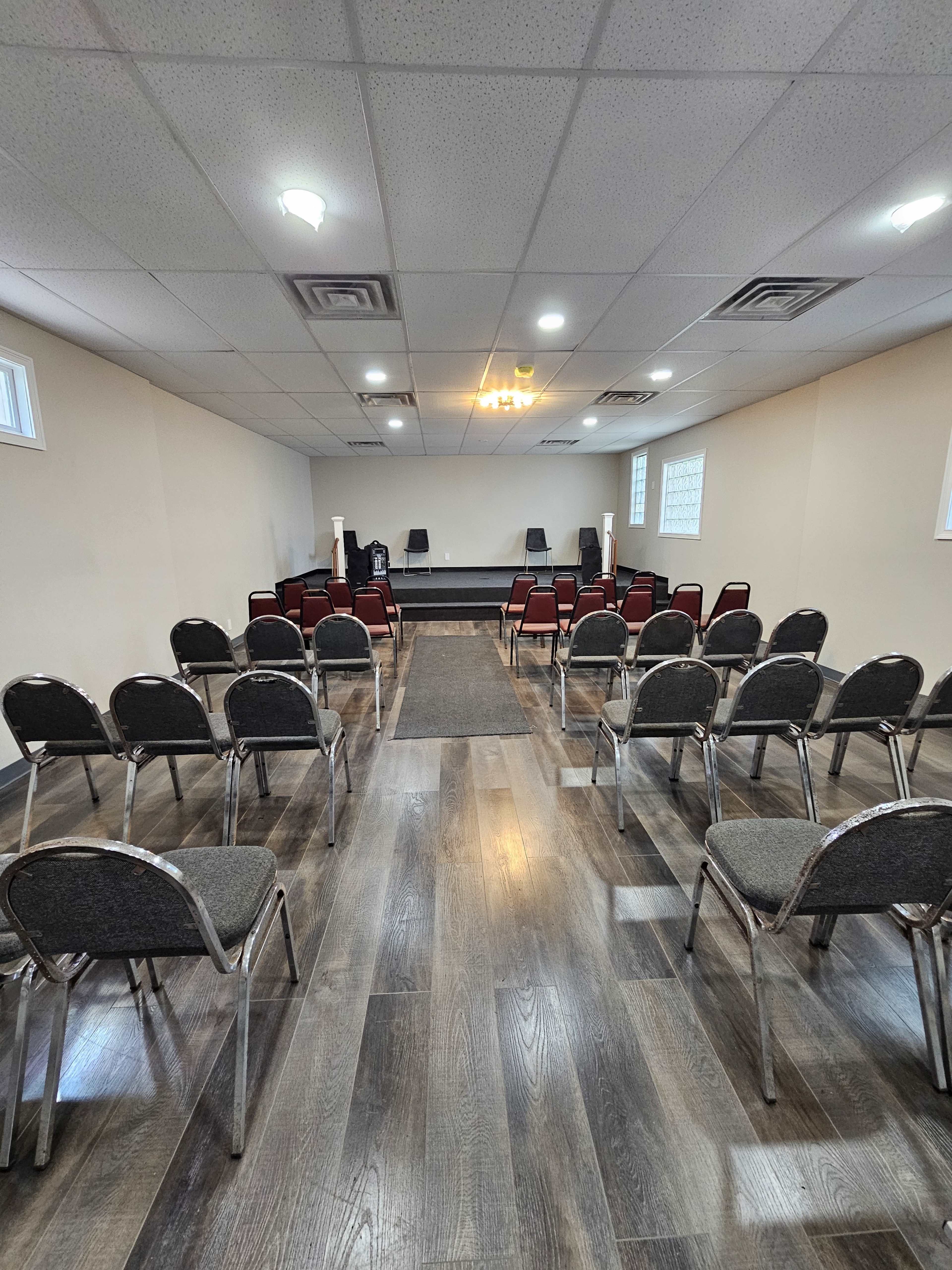 The image shows a sparse meeting room with rows of metal chairs facing a small stage at the front.