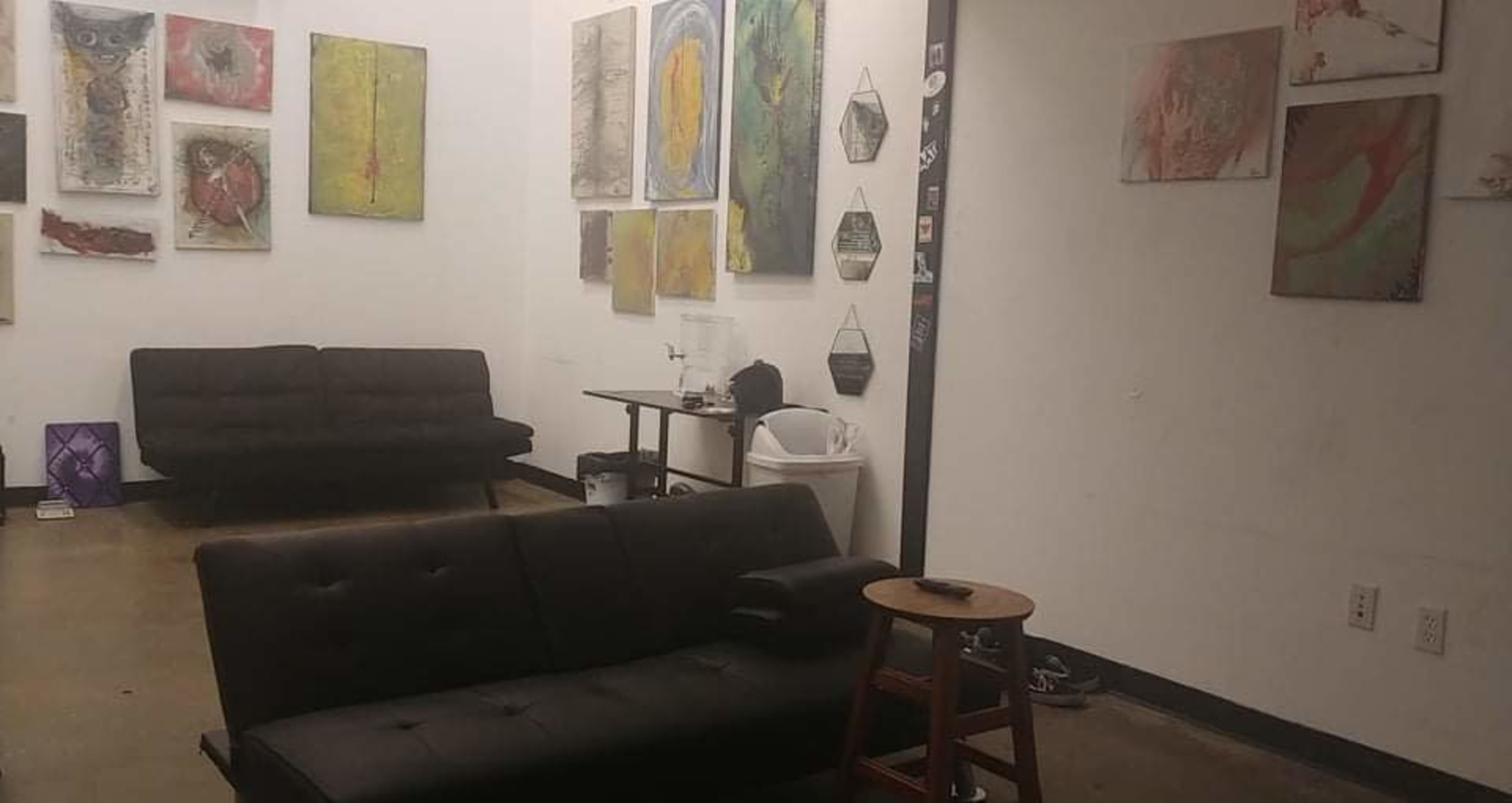 A minimalist art gallery featuring several abstract paintings on white walls, with two black sofas and a wooden stool in the foreground.